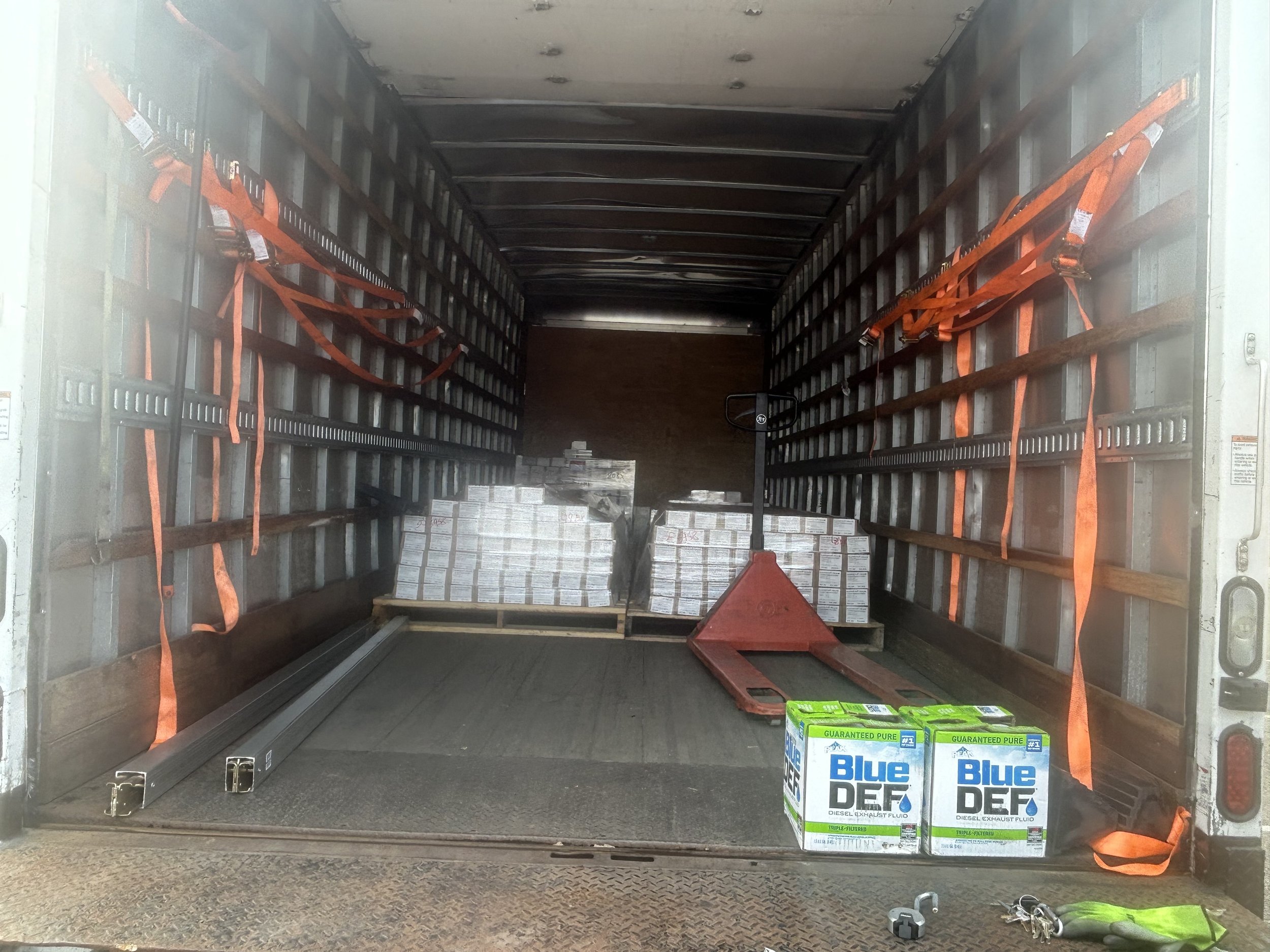 Empty truck cargo area with two boxes of Blue DEF diesel exhaust fluid and two boxes of engine parts, with palletized cargo in the background and various tools and straps inside.