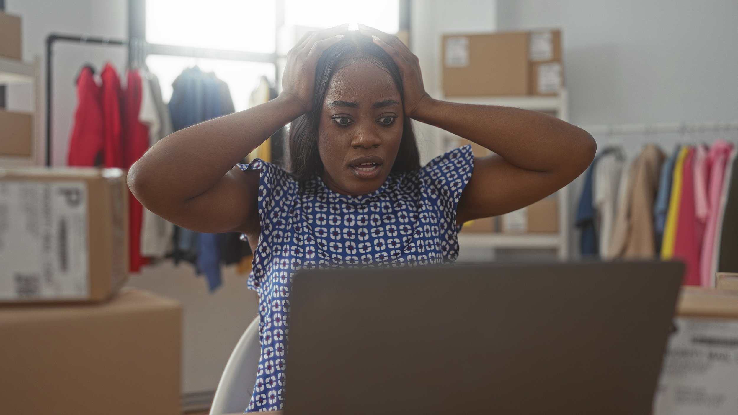 entreprenuer with hands on head overwhelmed in front of computer with retail shipments in the background
