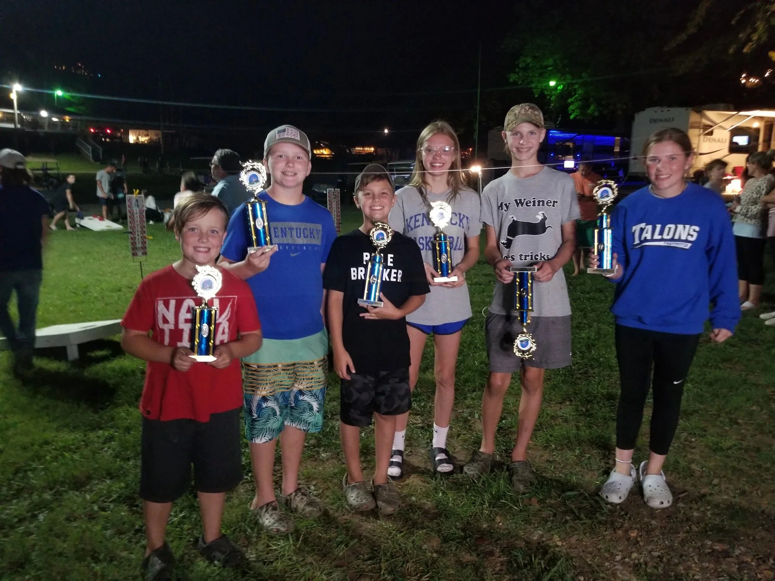 Seven children standing outdoors at night, holding trophies, smiling for the camera, with a crowd and tents in the background.