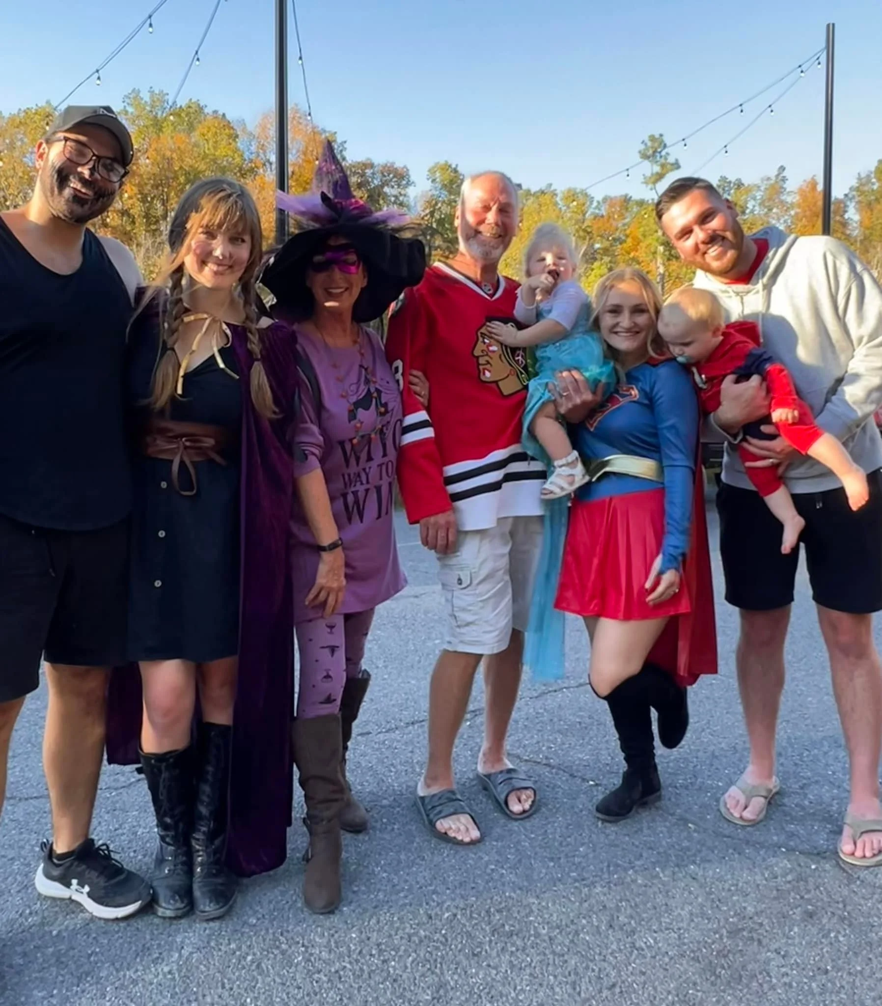 A group of nine people, including children, dressed in costumes, standing outside on a paved area with trees and string lights in the background.