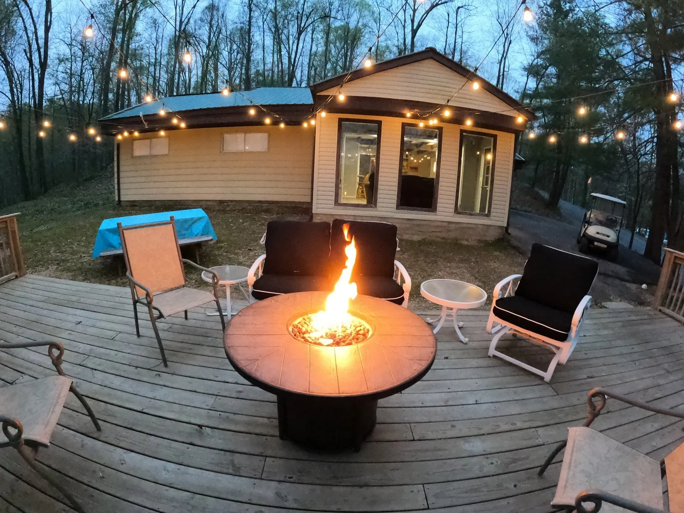 An outdoor wooden deck overlooking a wooded backyard during dusk, with string lights hanging overhead, a fire pit in the center, and patio chairs arranged around it.