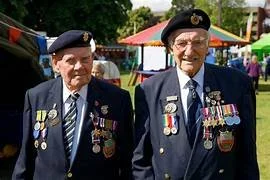 Two elderly men in uniform jackets decorated with medals, wearing berets, at an outdoor event with tents and people in the background.