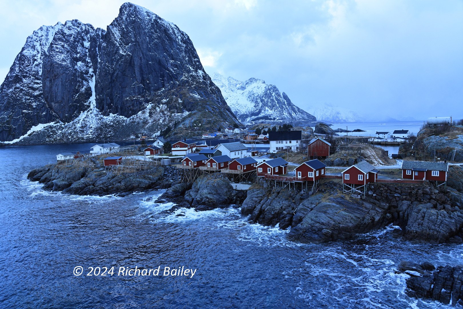 Red stilt houses with black roofs line the rocky shore of a Lofoten fjord below a snow-covered mountain.