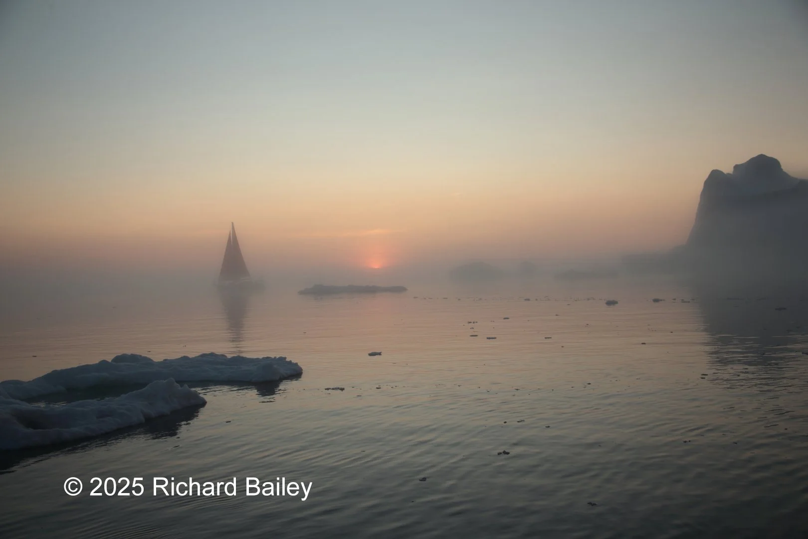 A sailboat and floating ice on calm water at sunset near a large rock formation, softened by fog off the Greenland coast.