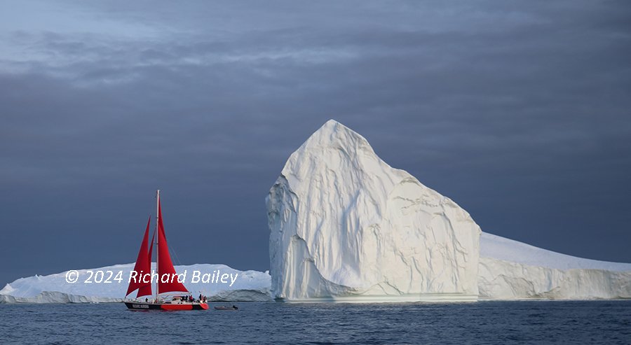 A red sailboat with red sails near a large iceberg against a cloudy sky.