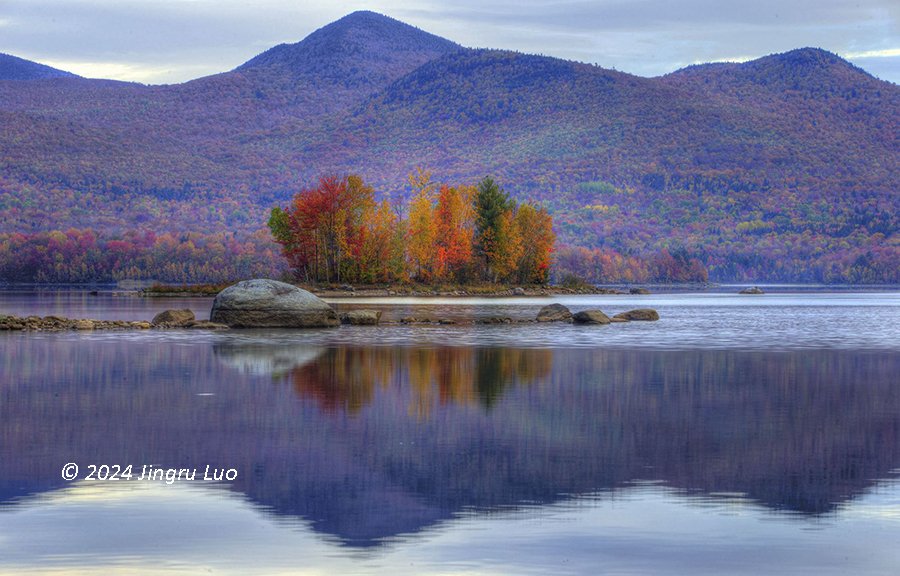 Small island with autumn-colored trees in the middle of a lake, surrounded by mountains under a cloudy sky.
