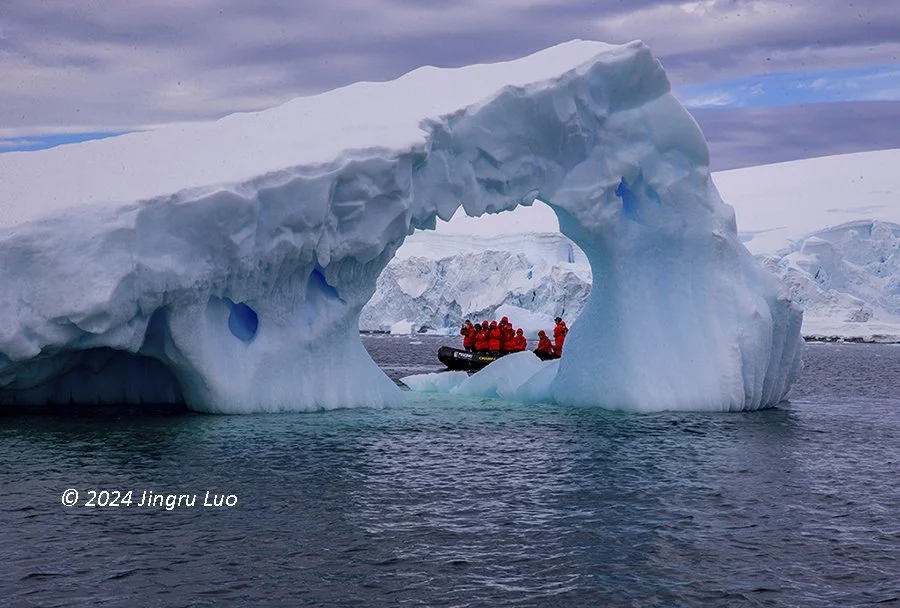 A Zodiac carrying passengers in orange jackets passes beneath a natural iceberg arch in polar waters.