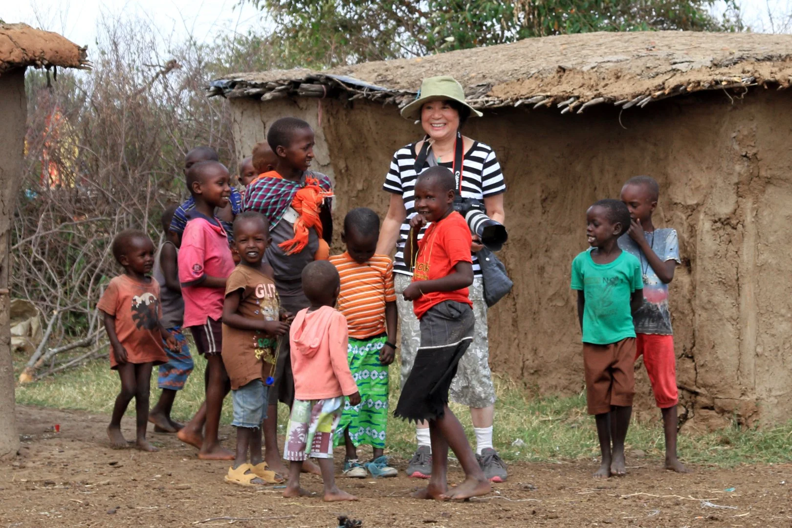 Photographer Jingru Luo stands smiling among a group of Maasai children outside traditional mud huts in rural Kenya.