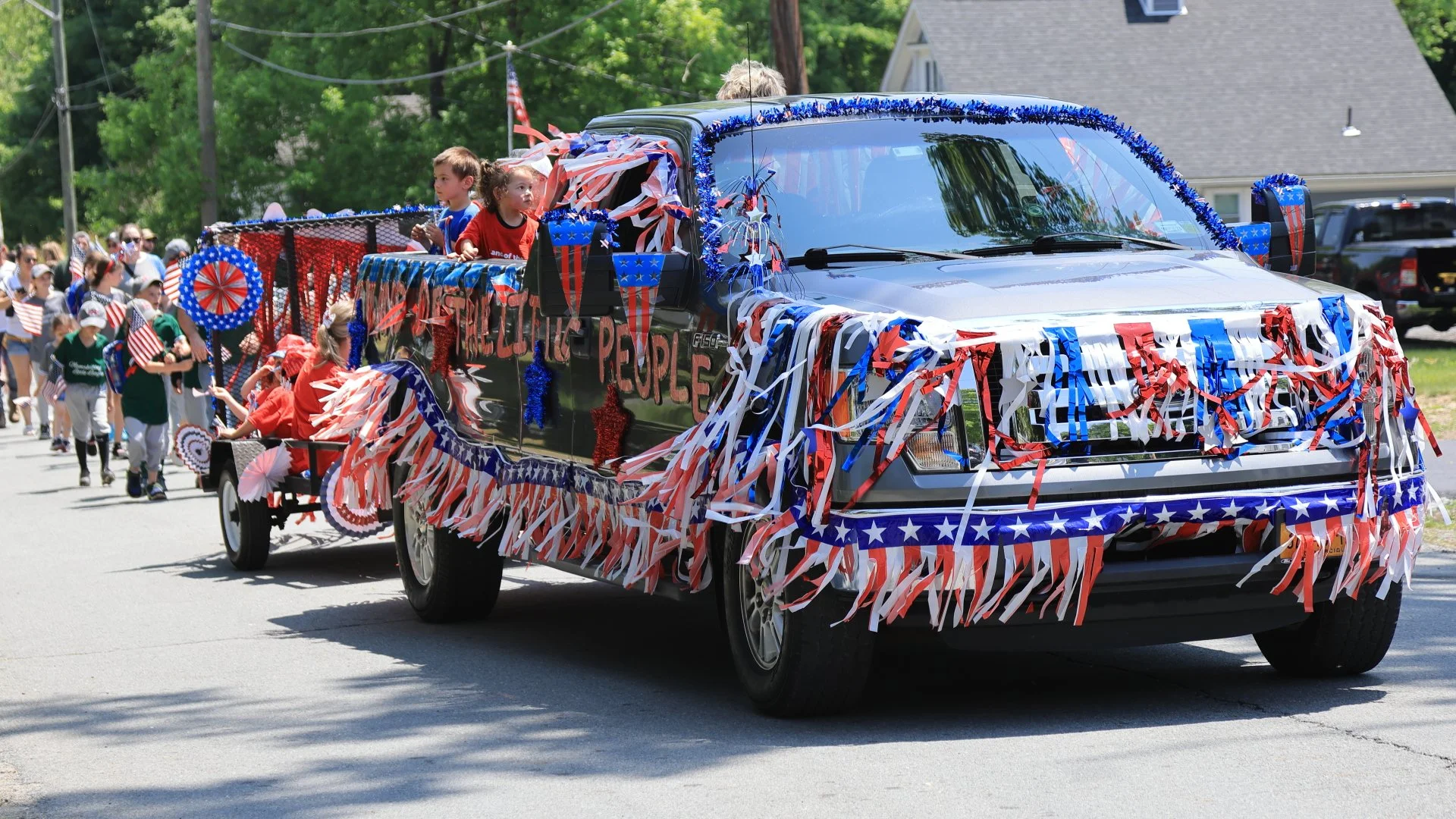 A decorated vehicle with red, white, and blue streamers and banners, participating in a parade on a sunny day. Children are riding on the vehicle, and a group of people, some holding American flag, walk along the parade route celebrating Memorial Day