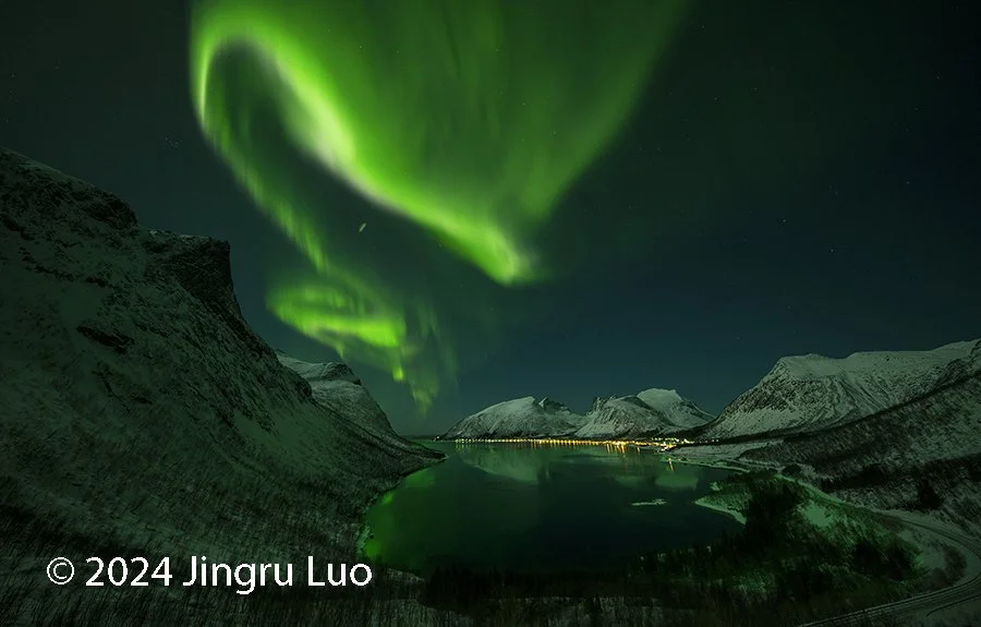 The aurora borealis curls into a dragon-like shape over snow-covered mountains and a calm river in Norway.
