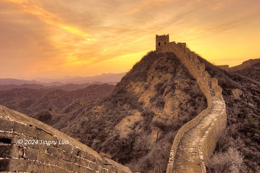 The Great Wall of China winds over hills at sunset with a watchtower silhouetted against a golden sky and distant mountains.