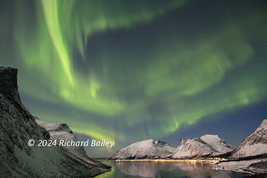 The aurora borealis dances above snow-covered mountains and water in Norway, with reflections on the surface.