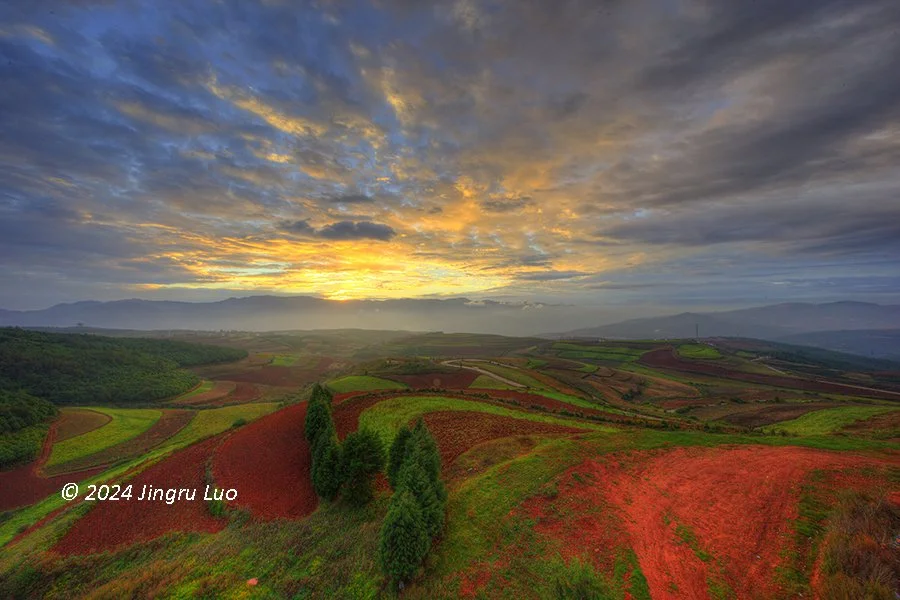 Sunrise over Dongchuan Red Land, China, showcasing vibrant red soil and lush green fields with a cloudy sky.