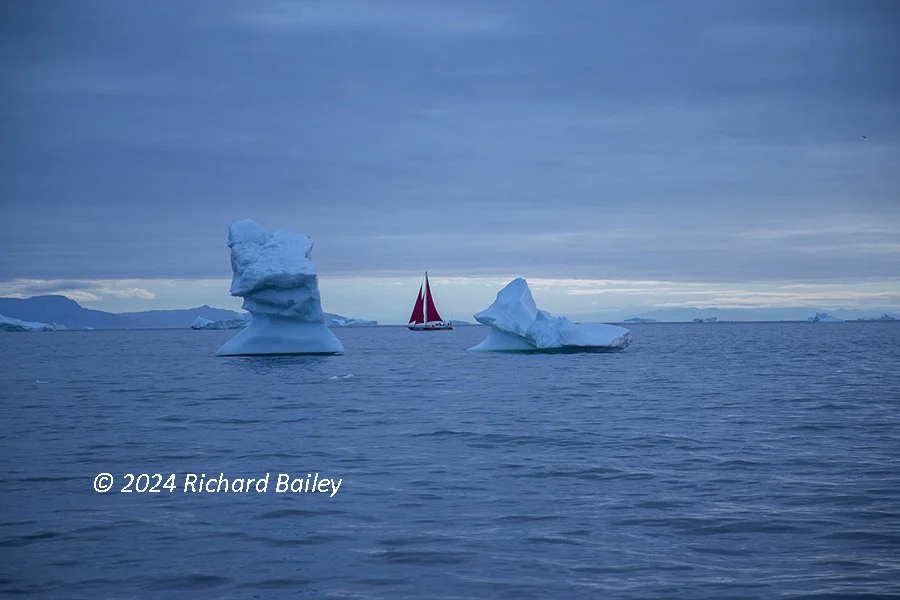 Sailboat navigating between icebergs in a cold, open sea under an overcast sky.