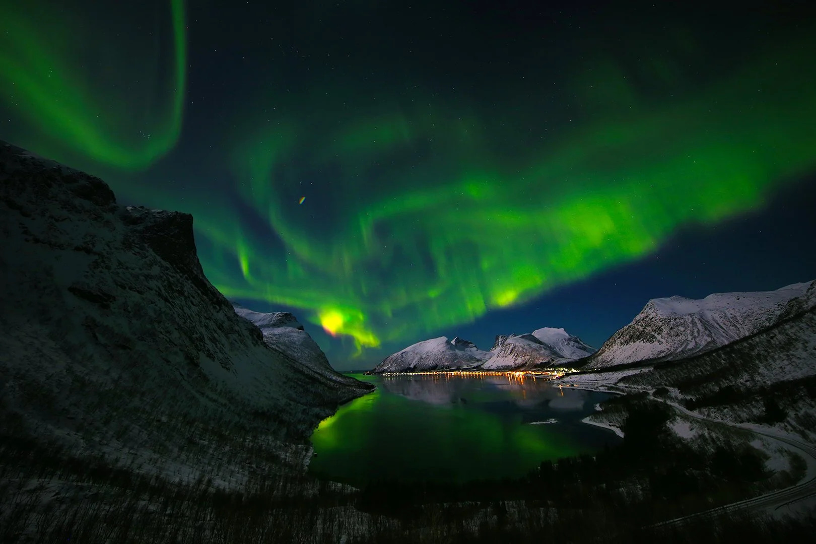 Northern lights over snow-covered mountains and a fjord at night, mirrored in the still water below.
