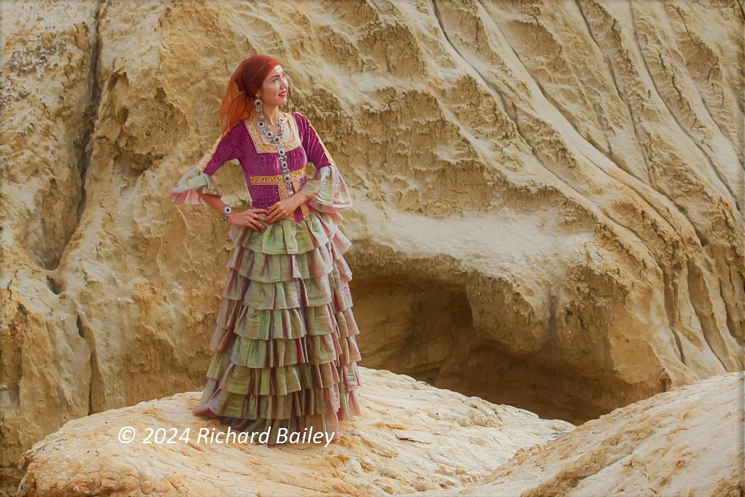 A model in traditional Xinjiang clothing stands on a rock formation against a backdrop of textured sandstone.