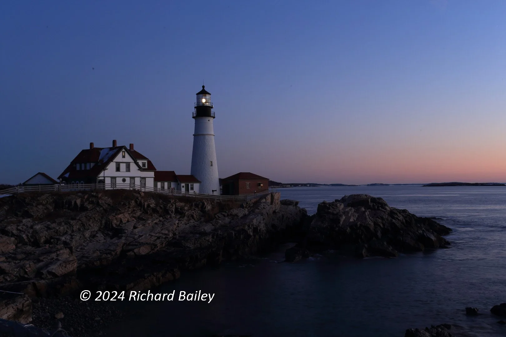 A lighthouse on a rocky coastline at dusk with a light beacon glowing, set against a clear sky with a gradient from blue to pink, and a calm ocean in the background.