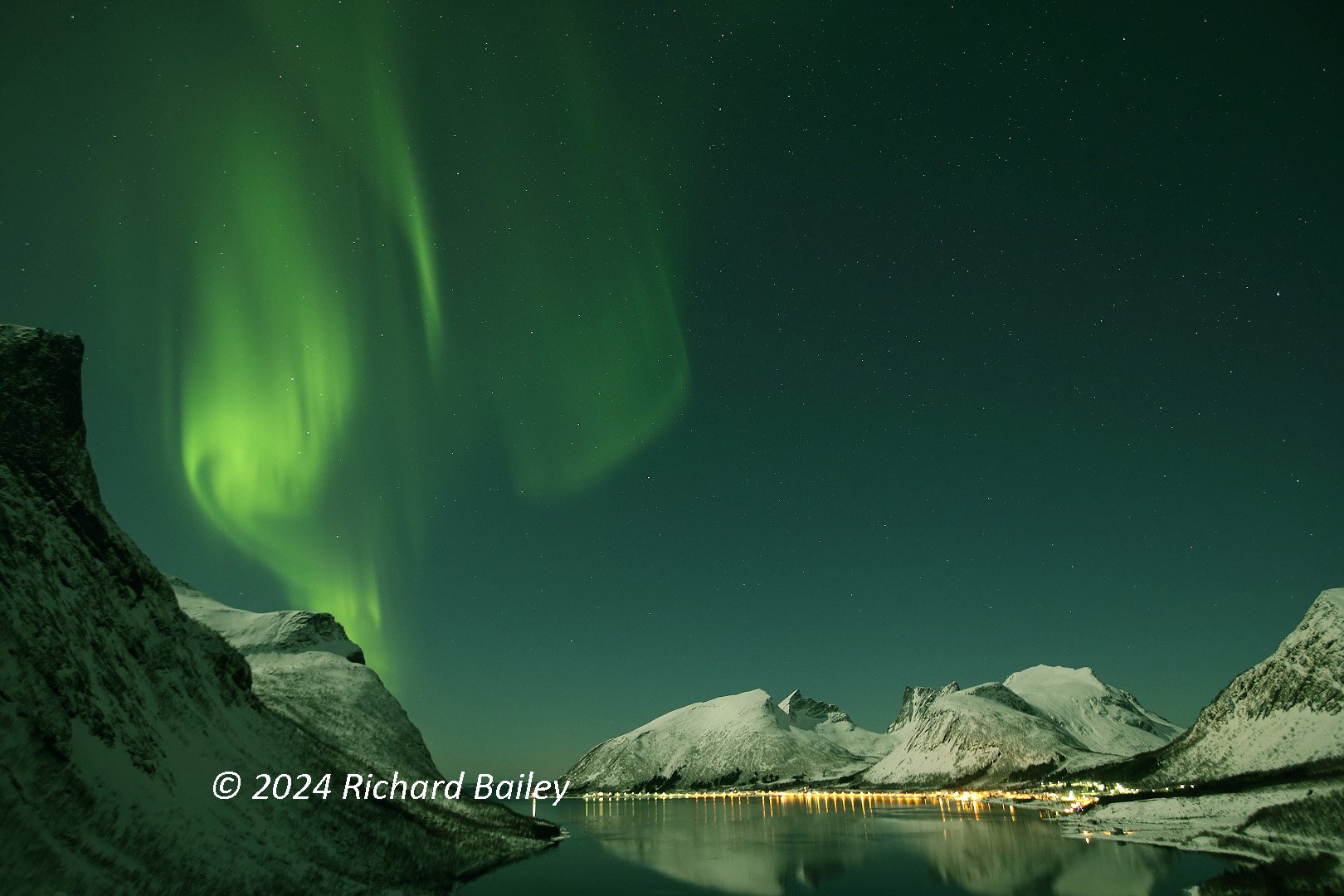 Swirling northern lights above snow-covered mountains and a fjord at night, with stars visible through the display.