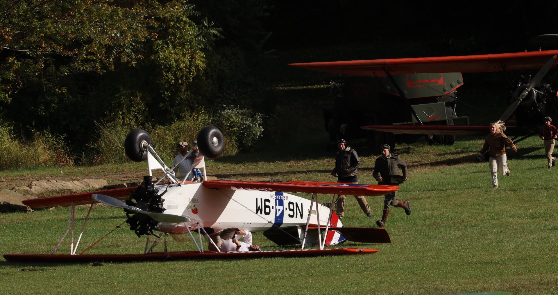 Vintage biplane flipped upside down on grass field, with people running nearby and trees in the background.