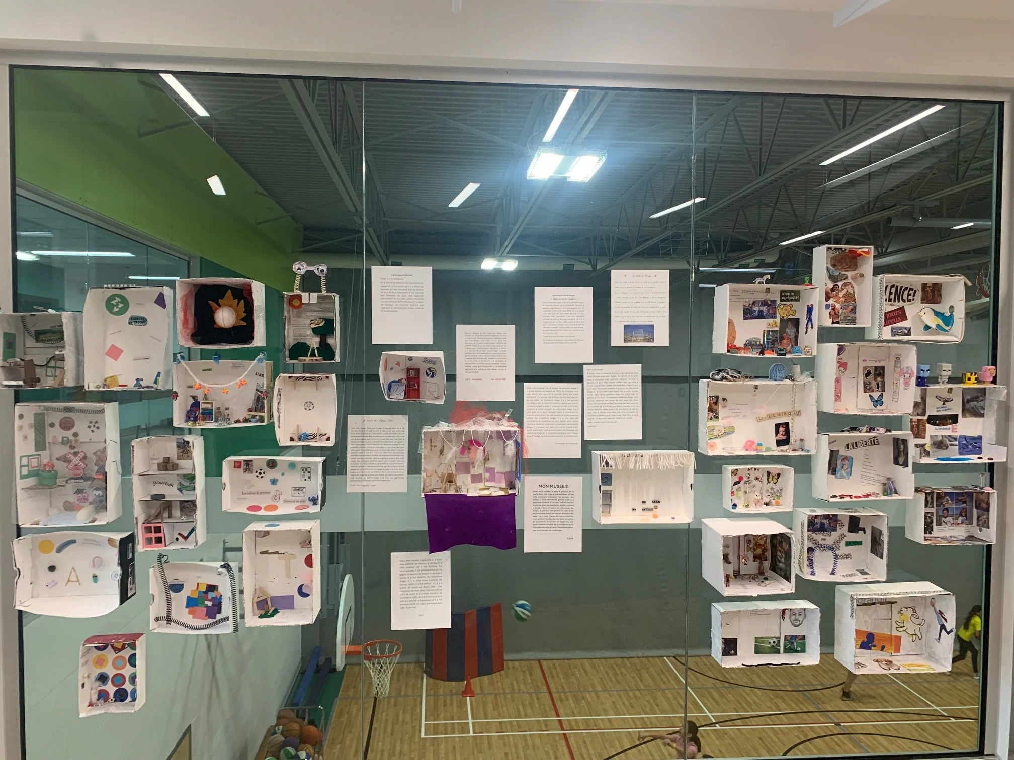 Exposition dans une salle de sport avec des boîtes en papier contenant diverses créations artistiques et textes affichés sur la vitre.