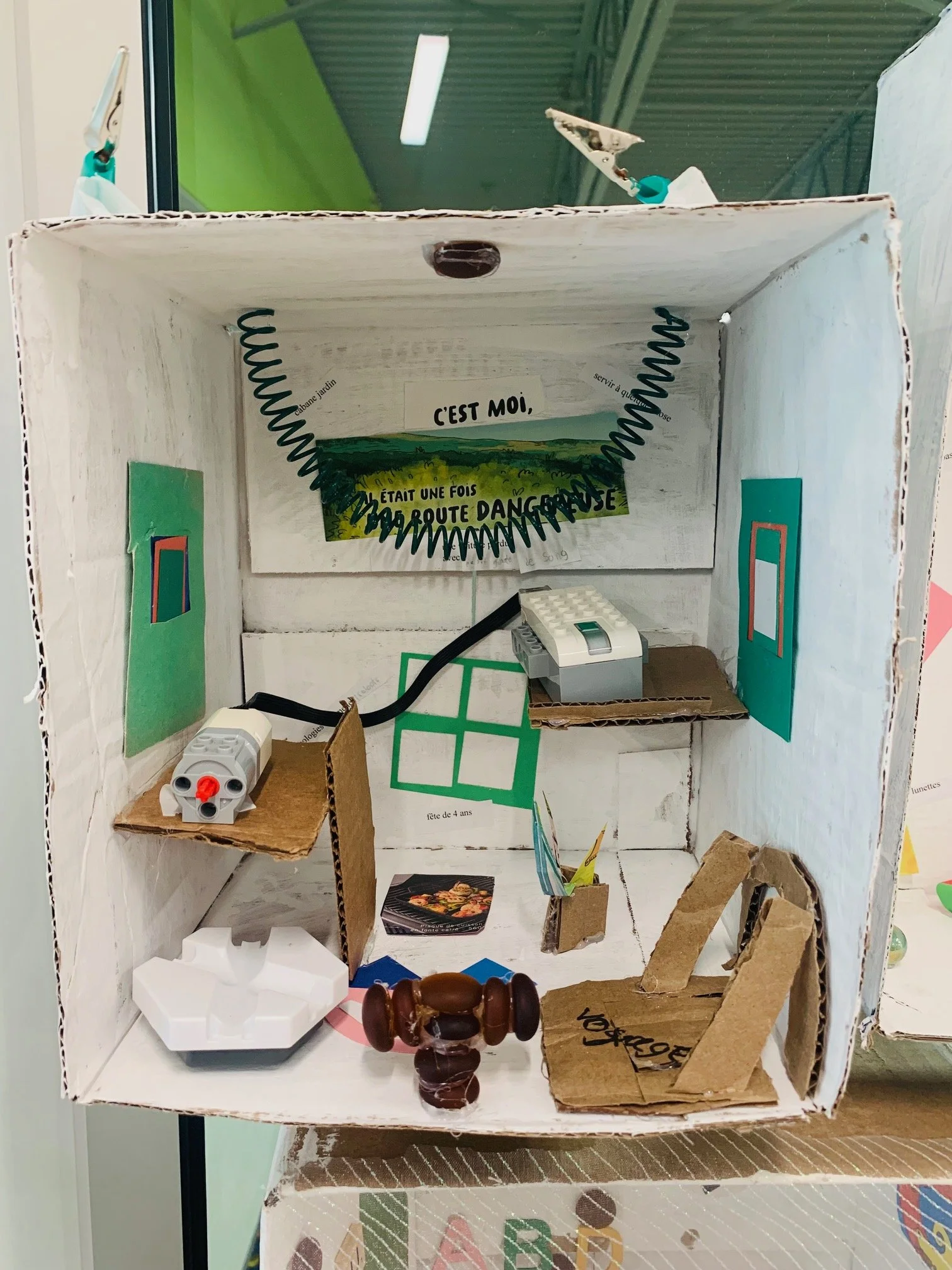 Une maquette de pièce réalisée avec une boîte en carton, comprenant des éléments en LEGO et du papier coloré, avec des messages écrits en français sur les murs.
