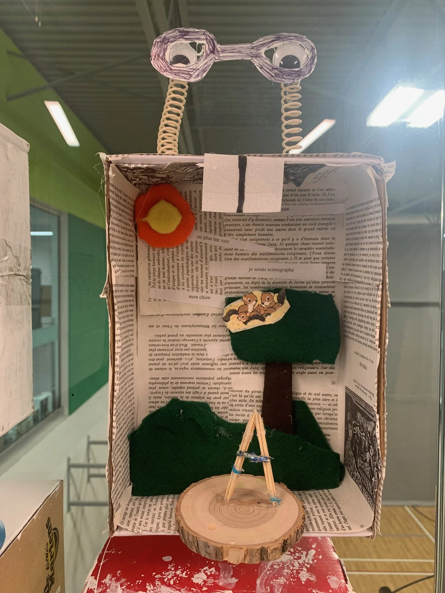 Une scène de théâtre miniature avec un arbre en feutrine, un cercle de bois représentant le sol et une figure de papiers représentant un buisson. Il y a aussi des éléments en feutrine représentant un soleil et un ciel. La scène est dans une boîte ave