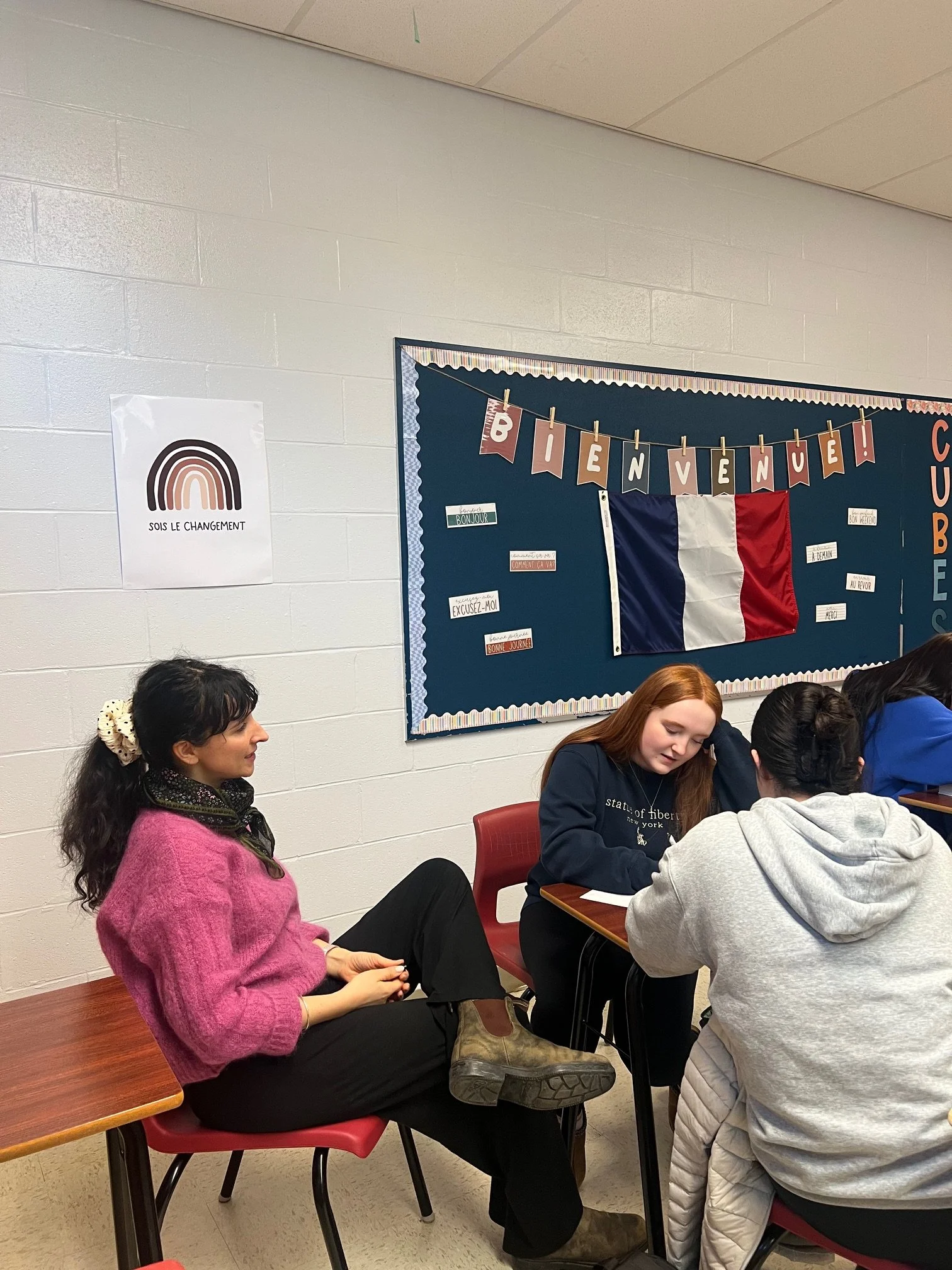 Des jeunes femmes discutent dans une salle de classe ou un espace communautaire avec un tableau derrière elles, décoré d'un drapeau français et d'une bannière 'Bienvenue'.