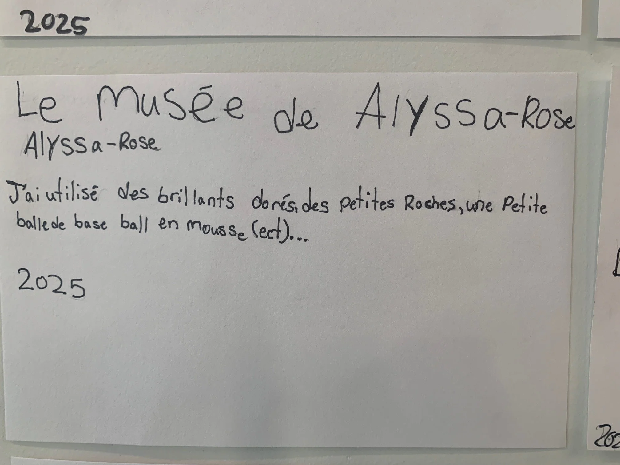 Carte blanche avec le titre "Le musée de Alyssa-Rose" et une description de son projet utilisant des petits cailloux, une petite boule de base ball en mousse, datée de 2025.