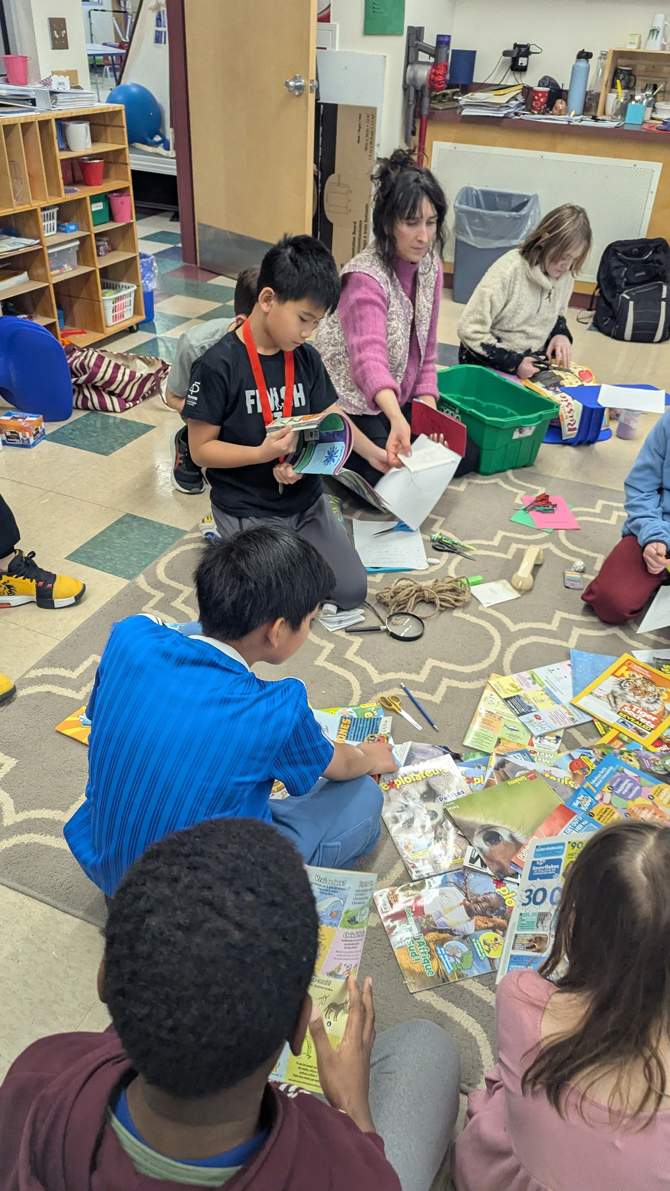 Des enfants et une adulte assis ou kneeling sur un tapis, entourés de magazines de science et nature, en train de lire ou d'explorer, dans une salle de classe ou une bibliothèque.