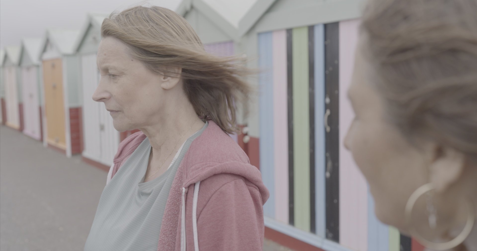 Two people standing near colorful beach huts, one in focus facing forward, wearing a pink hoodie.