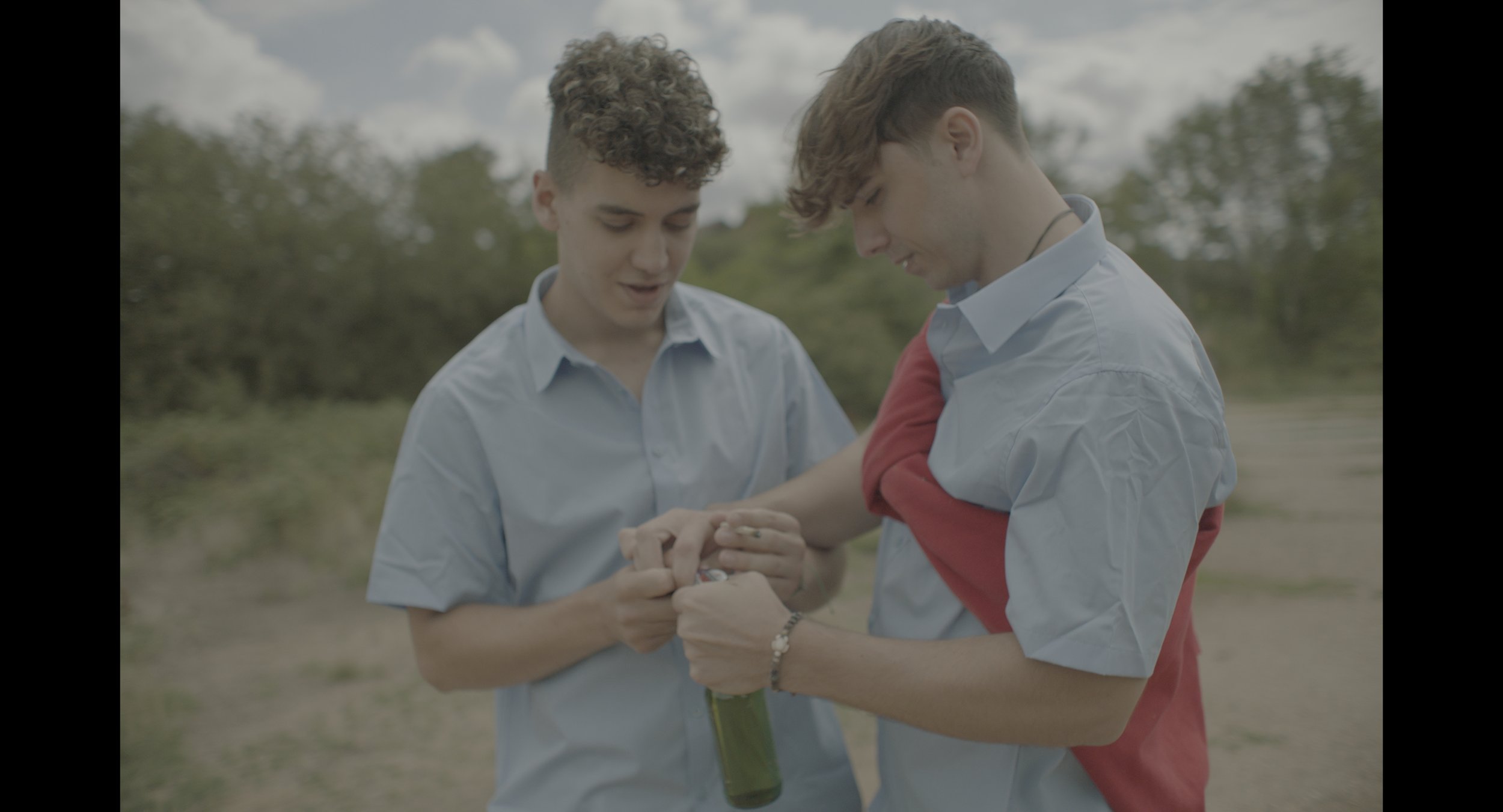 Two young men opening a bottle outdoors, wearing light blue shirts, with one wrapping a red garment around his shoulders.