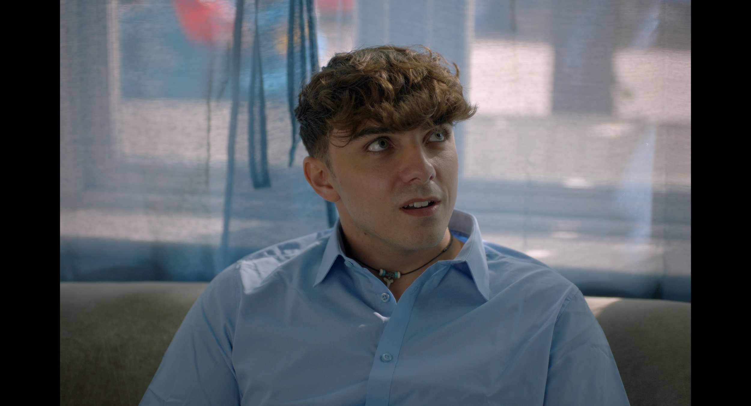 Young man in a light blue shirt looking surprised, sitting on a couch with a softly lit background.