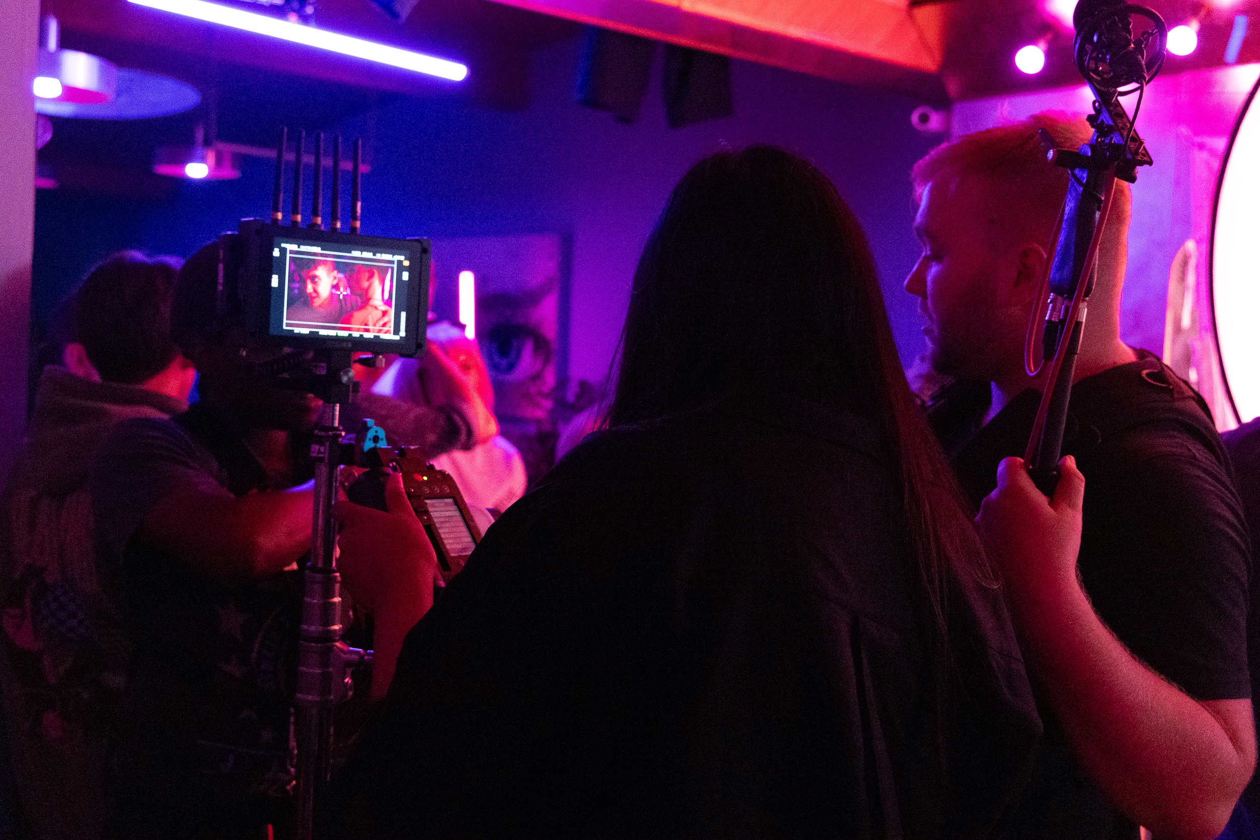 Behind-the-scenes view of a film set with crew members and equipment in dim lighting, featuring a camera screen displaying a scene.