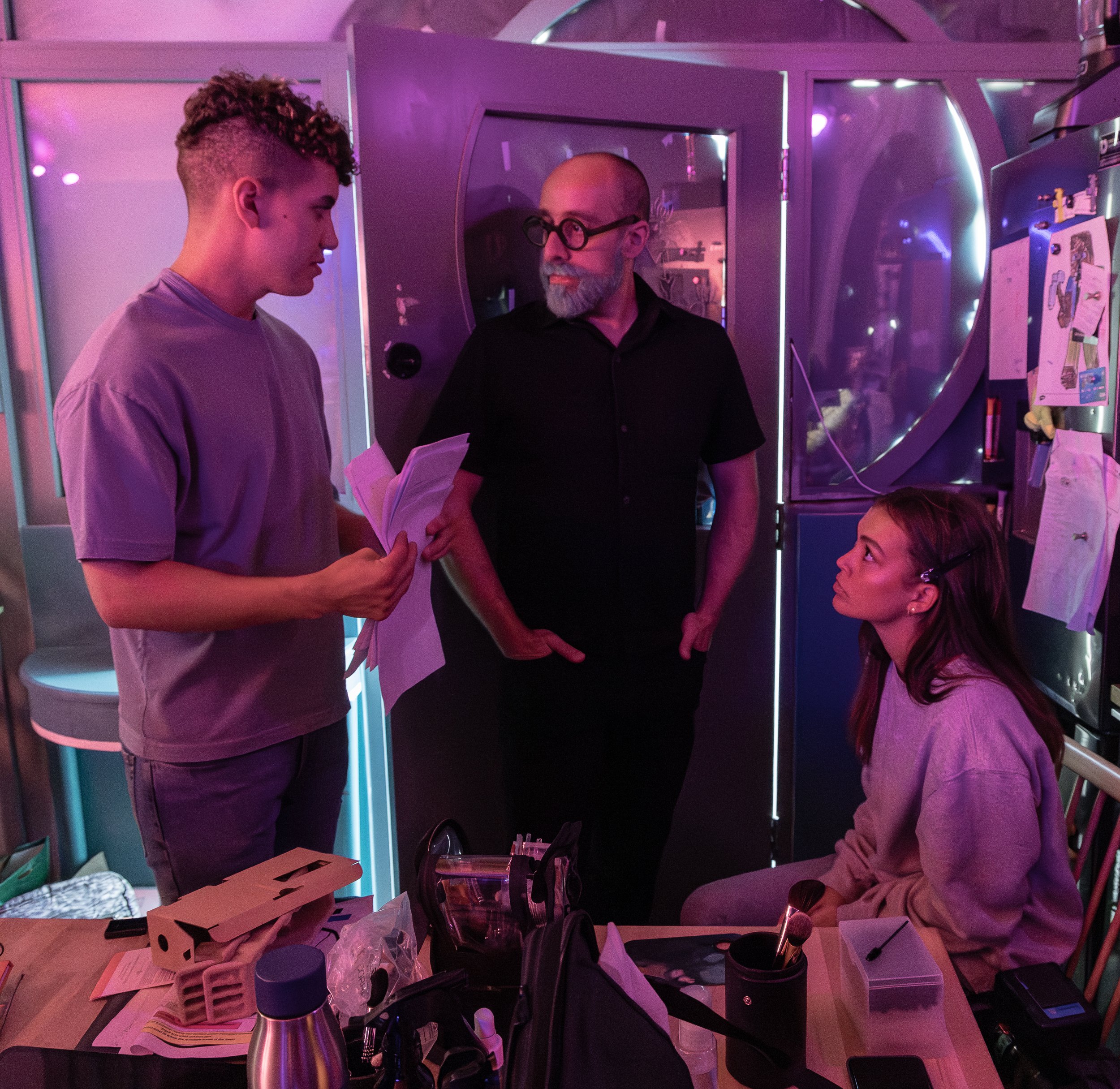 Three people in a purple-lit room, one standing and holding papers, one seated with hair clips, and one with a beard and glasses, near a cluttered table with supplies.