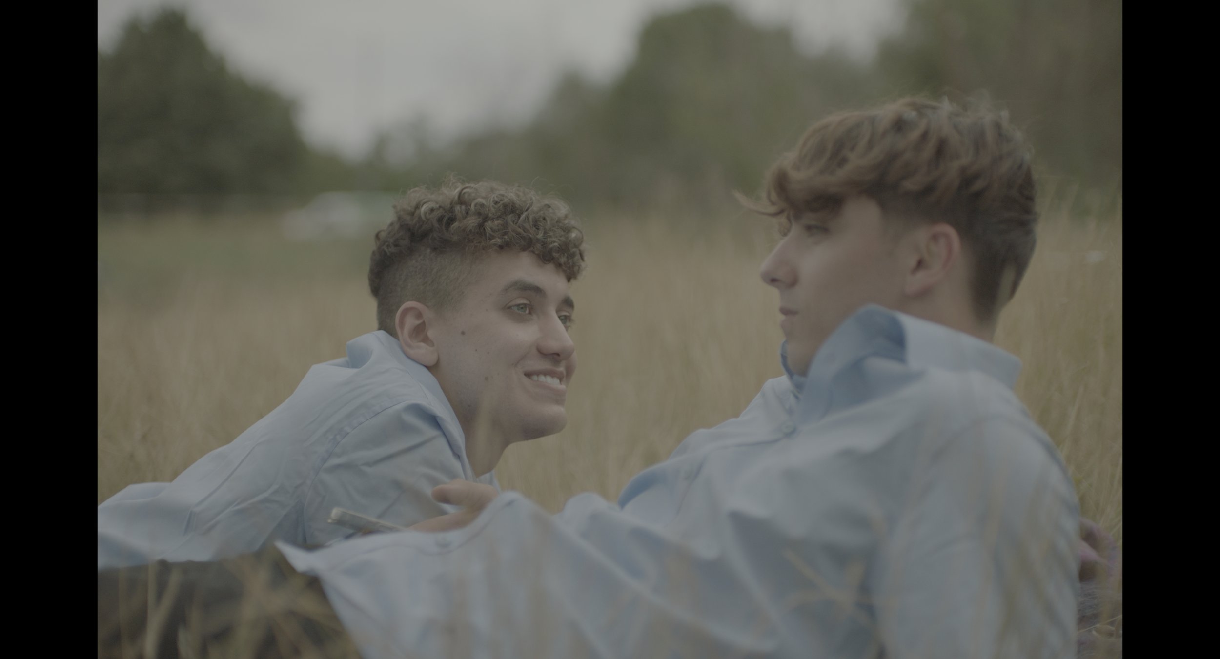 Two young men lie in a grassy field, wearing blue shirts. One is smiling at the other.