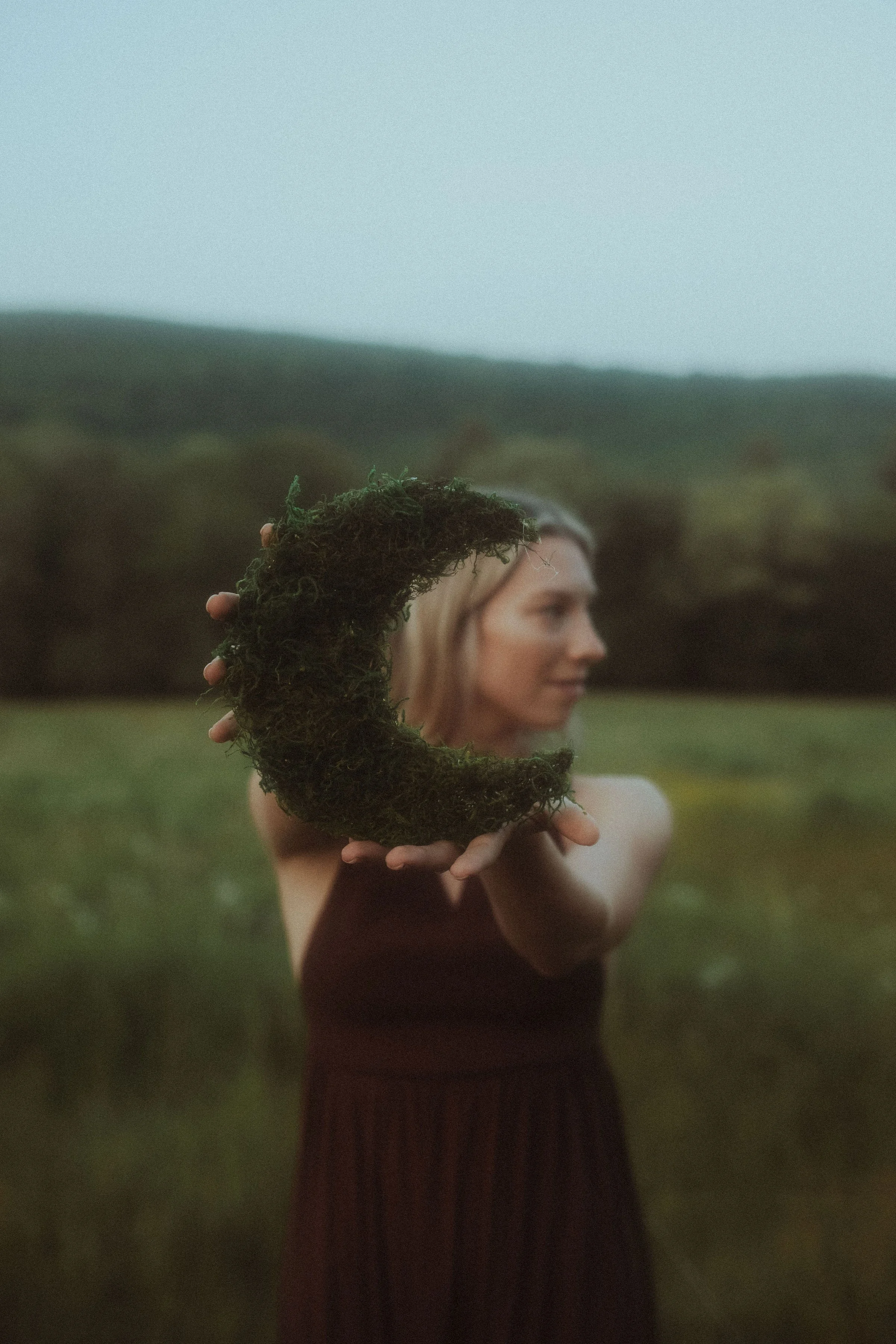 A woman standing outdoors holding a moss moon sculpture in front of her face with a forested hillside in the background.