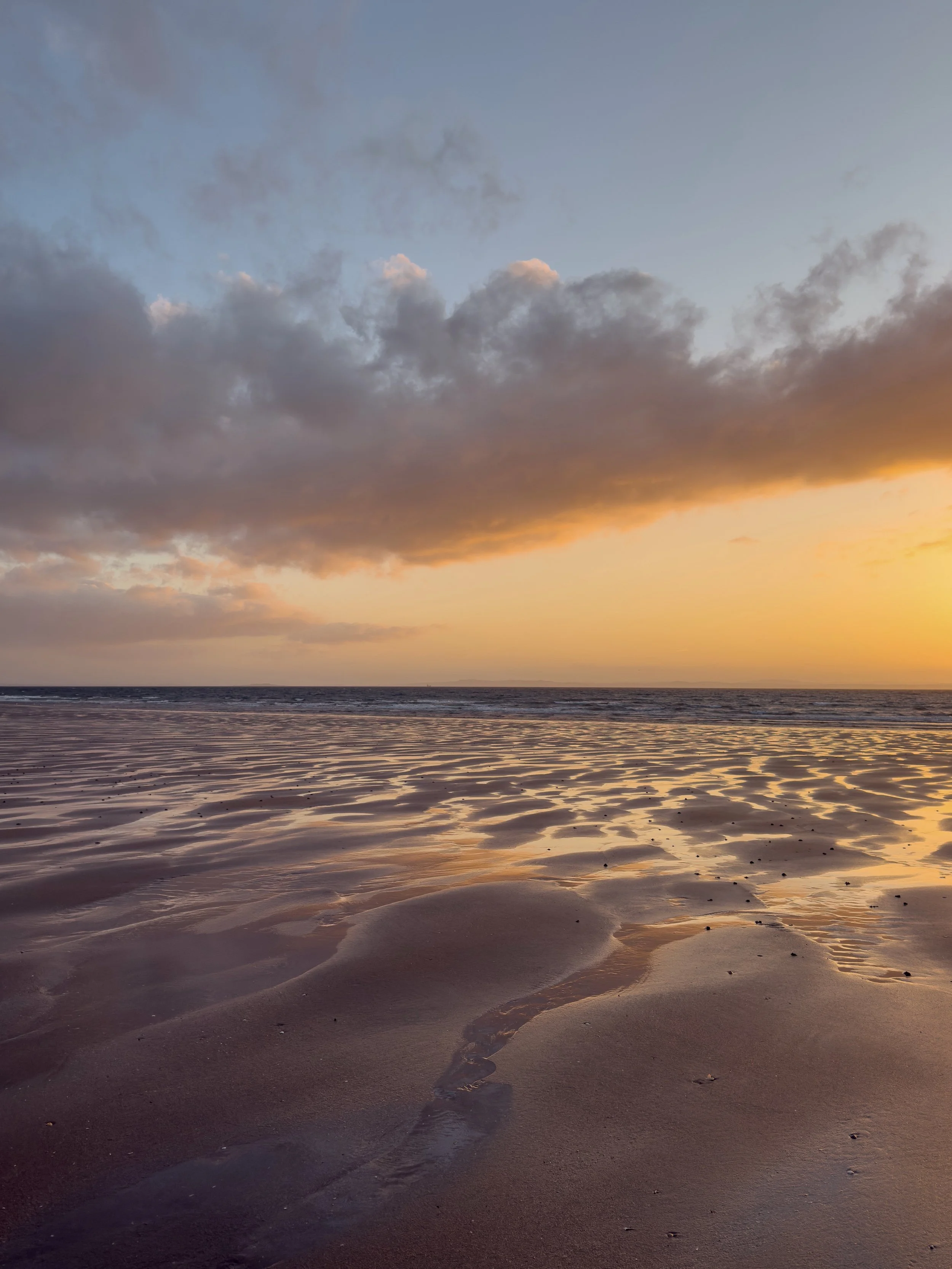 A beach at low tide, with the setting sun glowing across the wet sand and in the clouds above.