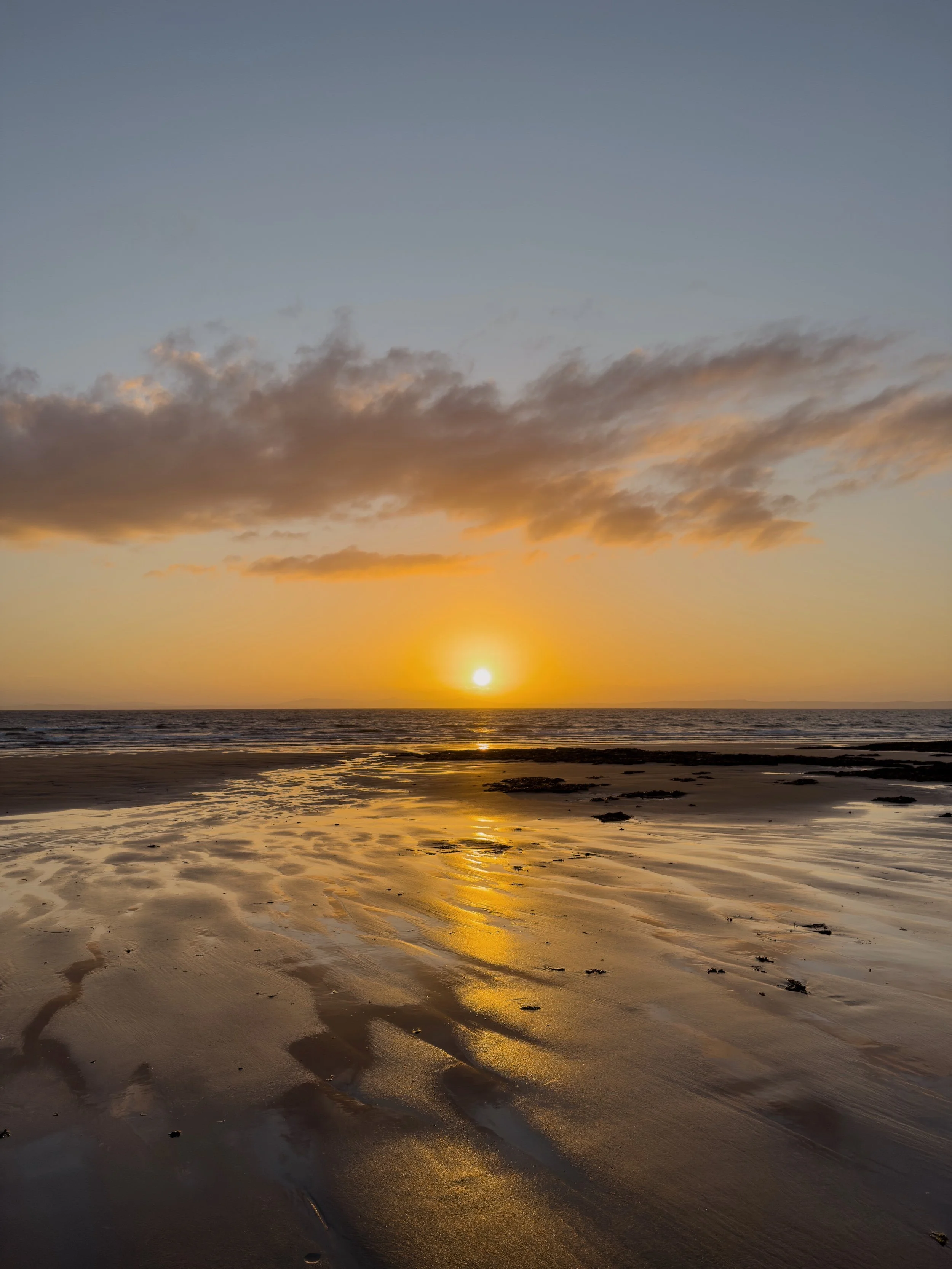 A beach at low tide, with the setting sun glowing across the wet sand and in the clouds above. The sun sits just above the horizon.
