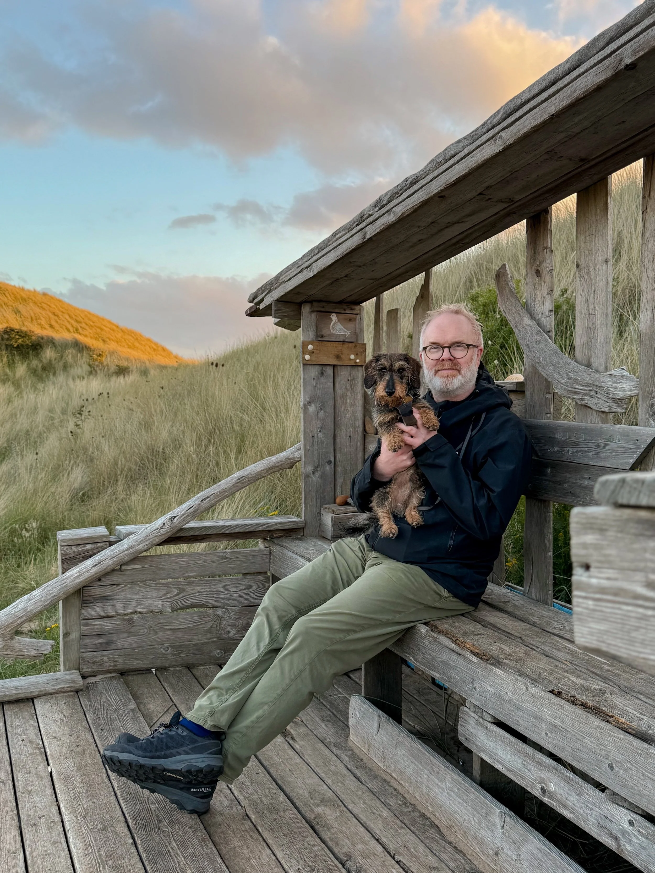 A man and a wirehaired dachshund sit at a driftwood shelter tucked behind the dunes, with glowing light on the dunes in the background and in the clouds above.