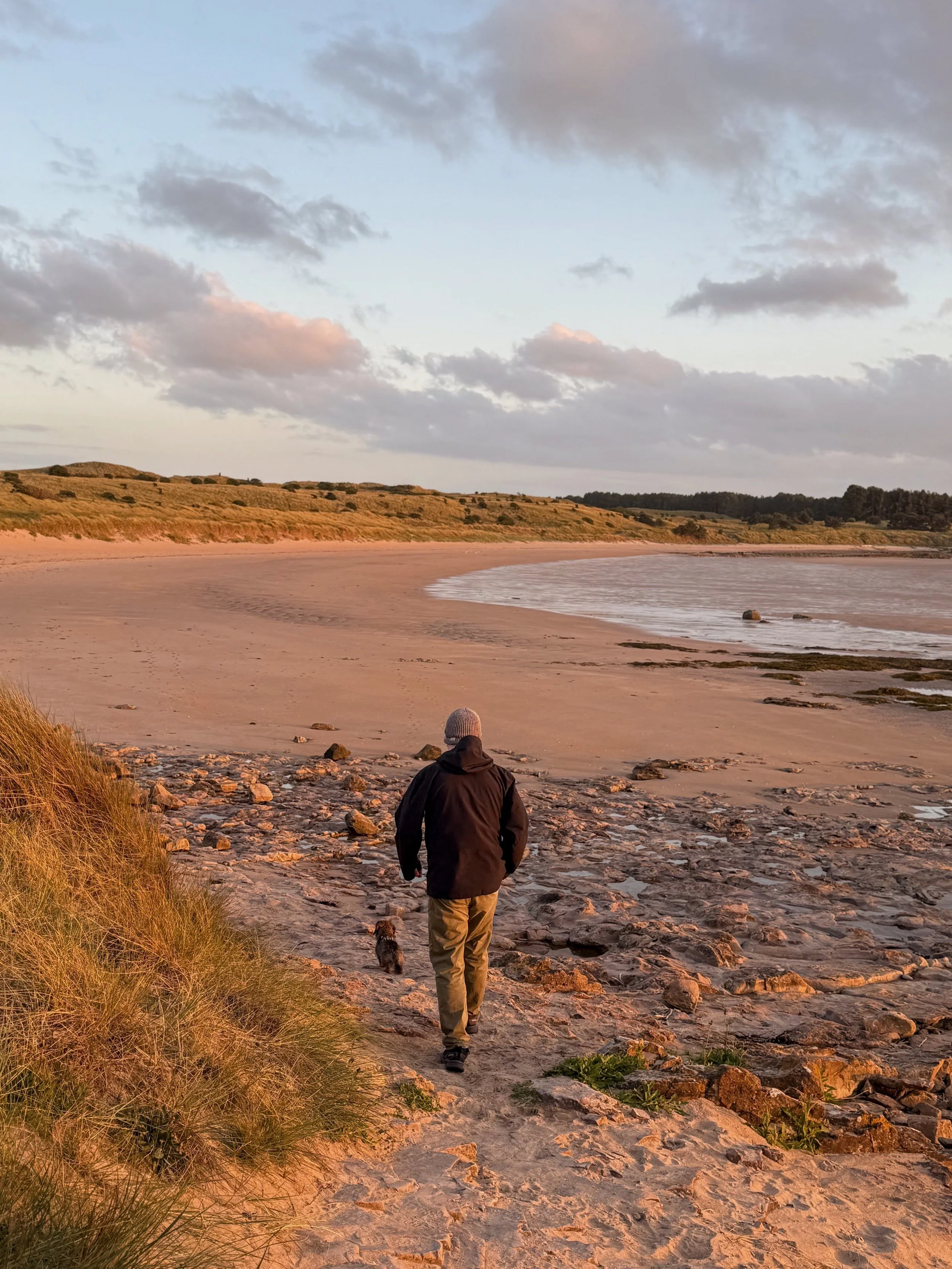 A man walks down onto a beach at low tide, with the dunes sweeping in a curve behind the shore. The sun is setting, casting a warm glow across the scene. A miniature wirehaired dachshund walks beside the man.
