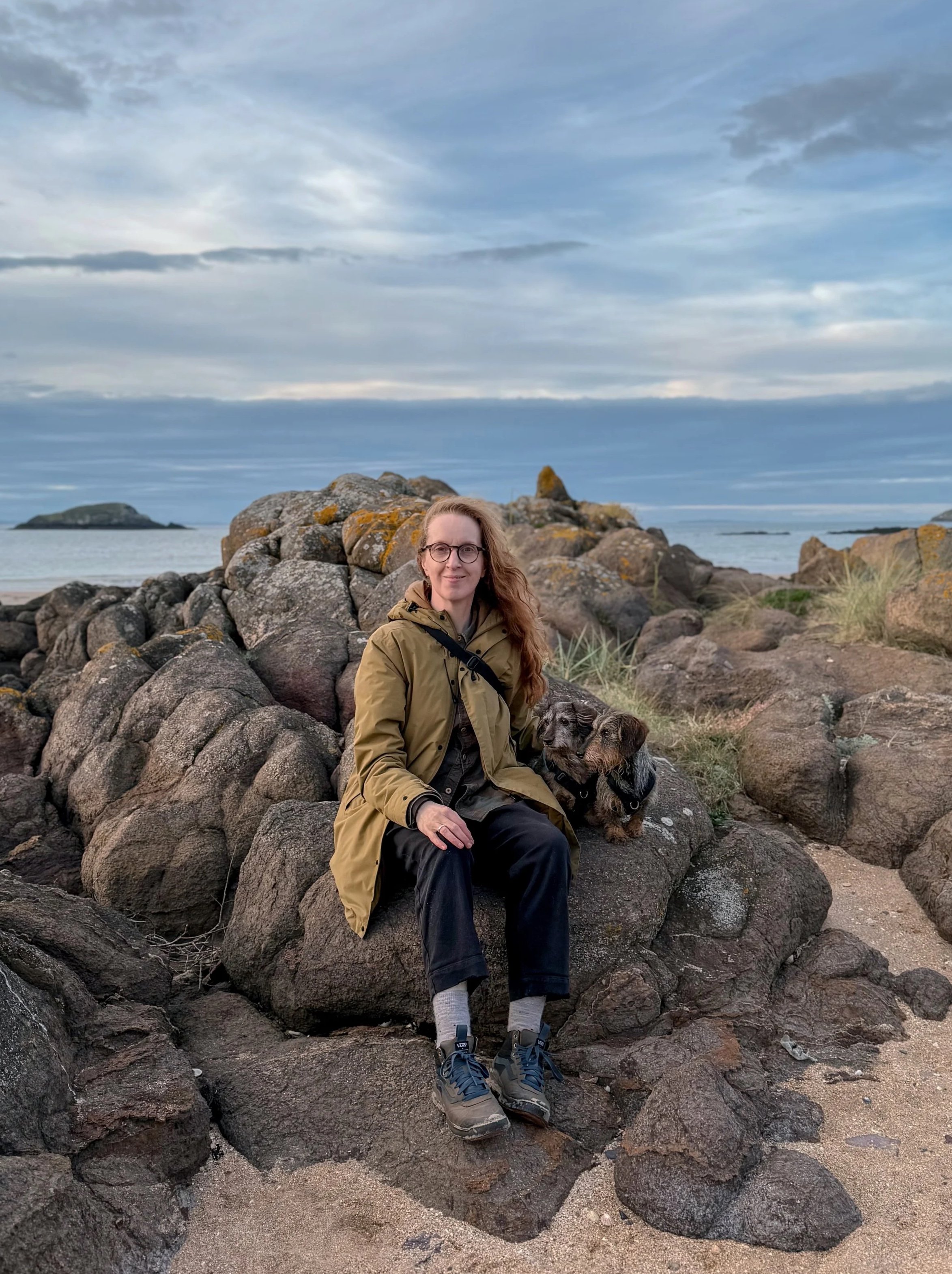 A woman with glasses and long red hair sitting on rocks near the beach, accompanied by two wirehaired dachshunds The woman is wearing a lichen coat, black trousers, and hiking shoes. The background features a cloudy sky, water, and distant islands.