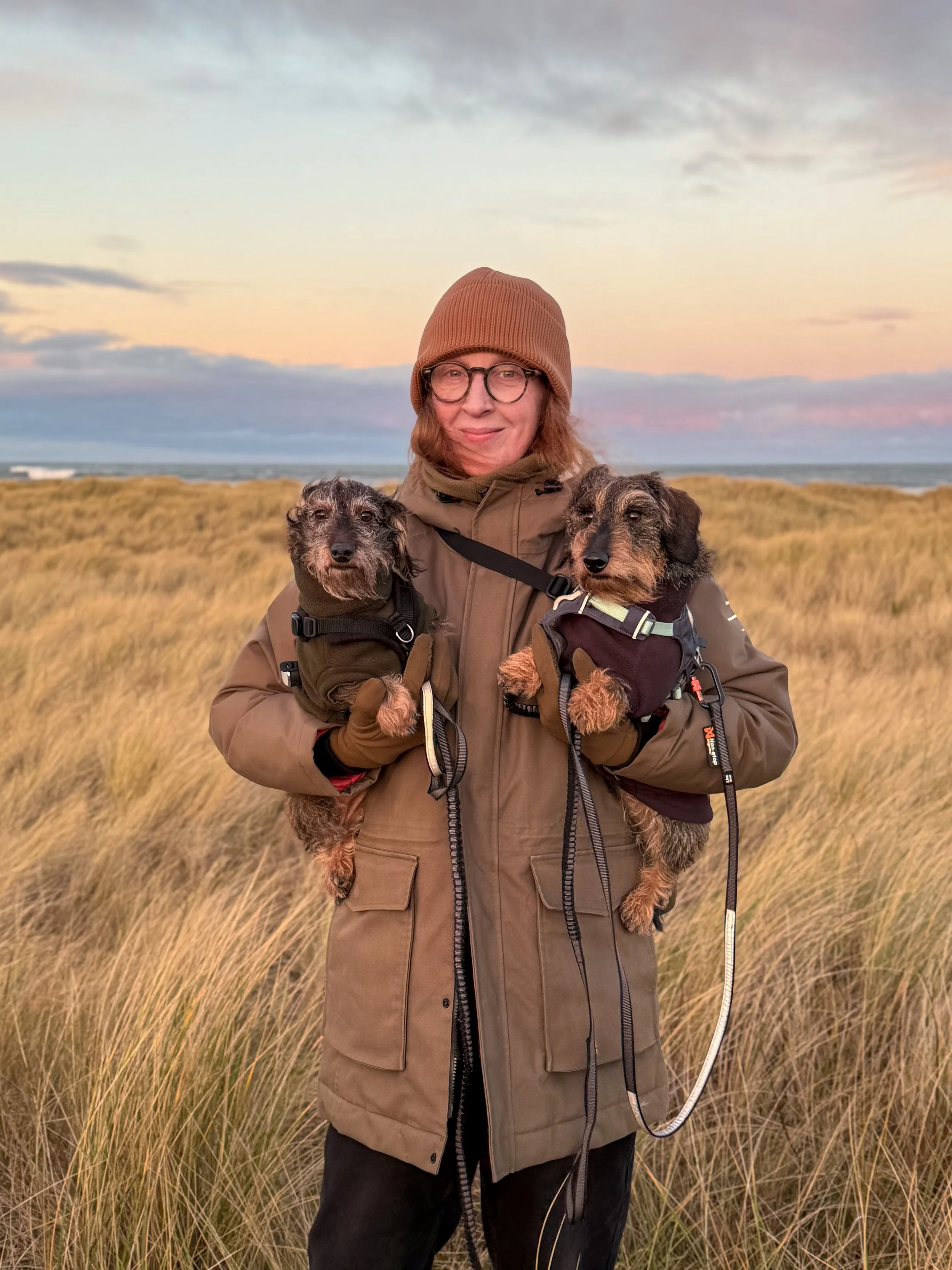 A woman with glasses and red hair holding two dachshunds in the dunes.