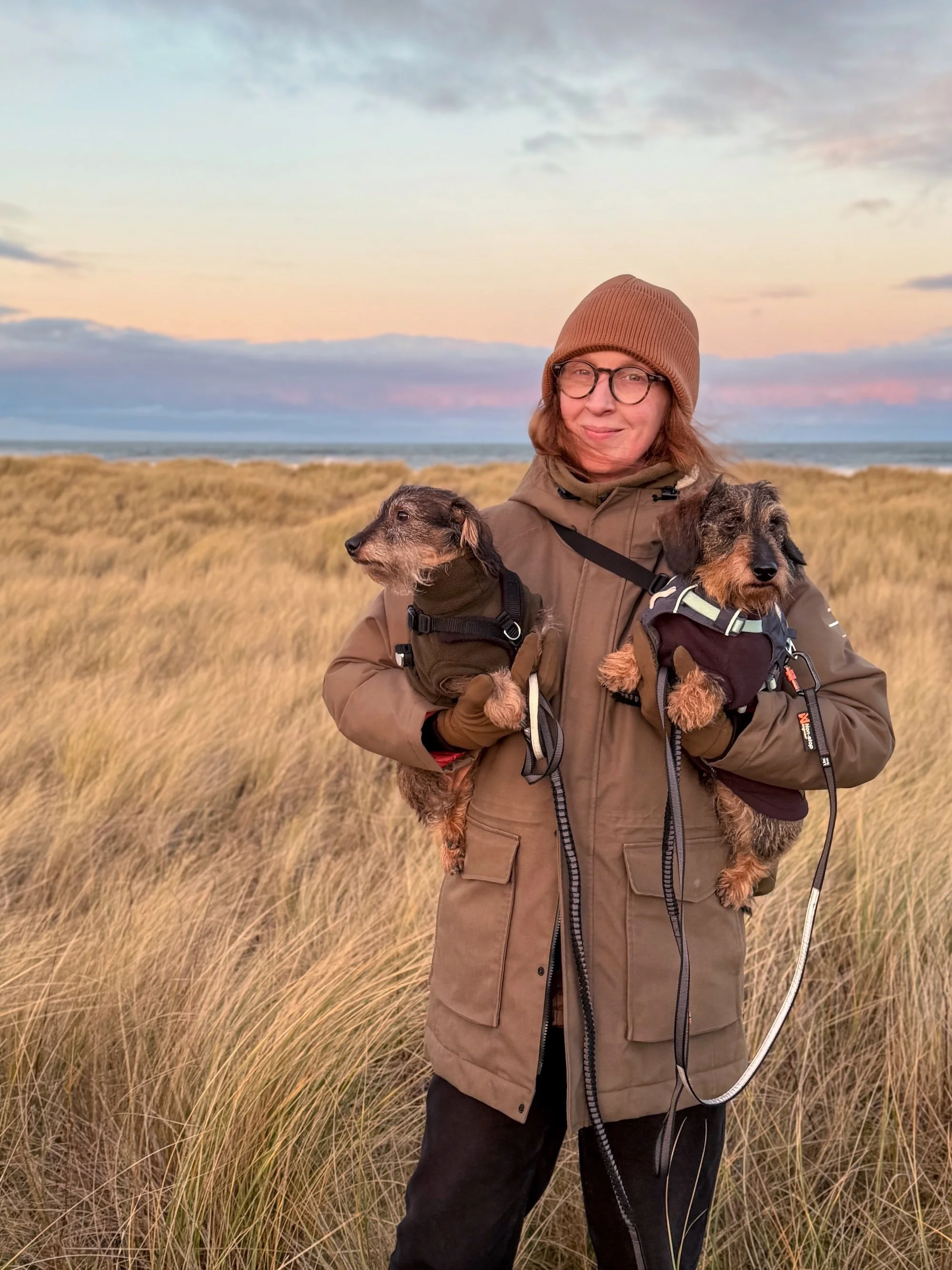 A woman with glasses and red hair holding two dachshunds in the dunes.