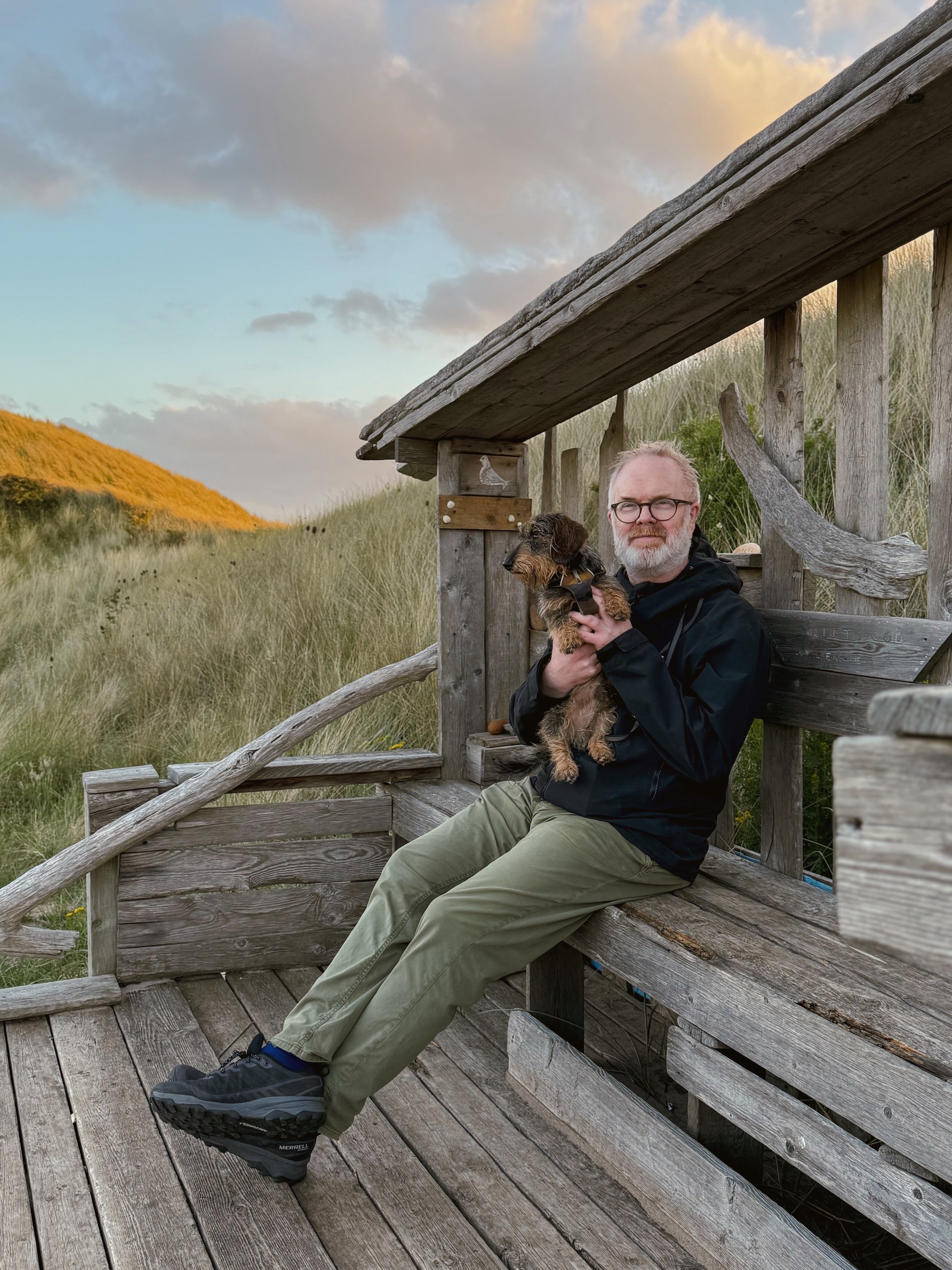 A man holds a wirehaired dachshund, sitting in a driftwood shelter tucked behind the dunes. The man is smiling to the camera. There is sunlight glowing on the dune grasses in the distance.