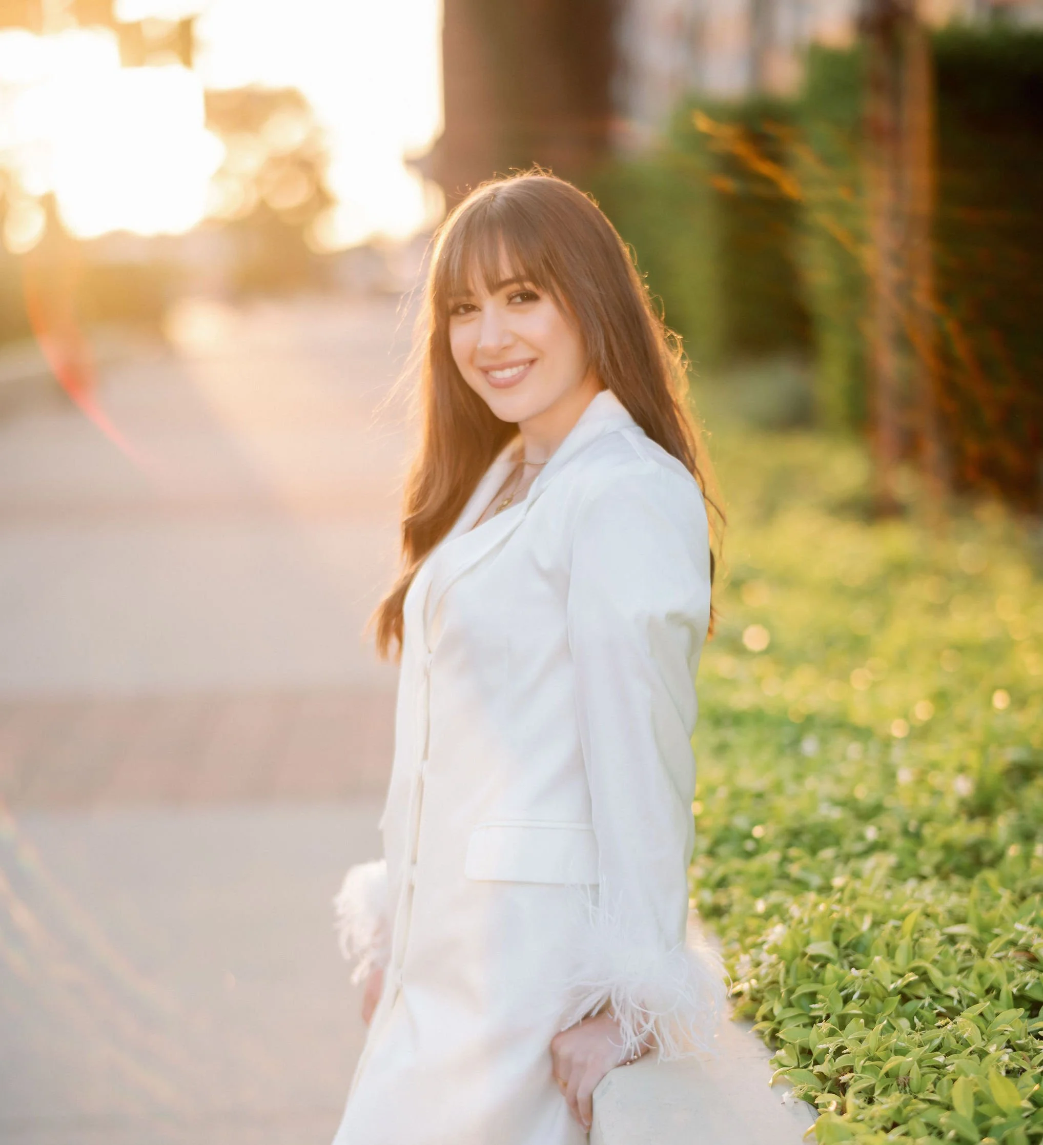 A young woman with long brown hair smiling at the camera, wearing a white blazer with feathered cuffs, standing outdoors in front of a sunlit background with greenery.