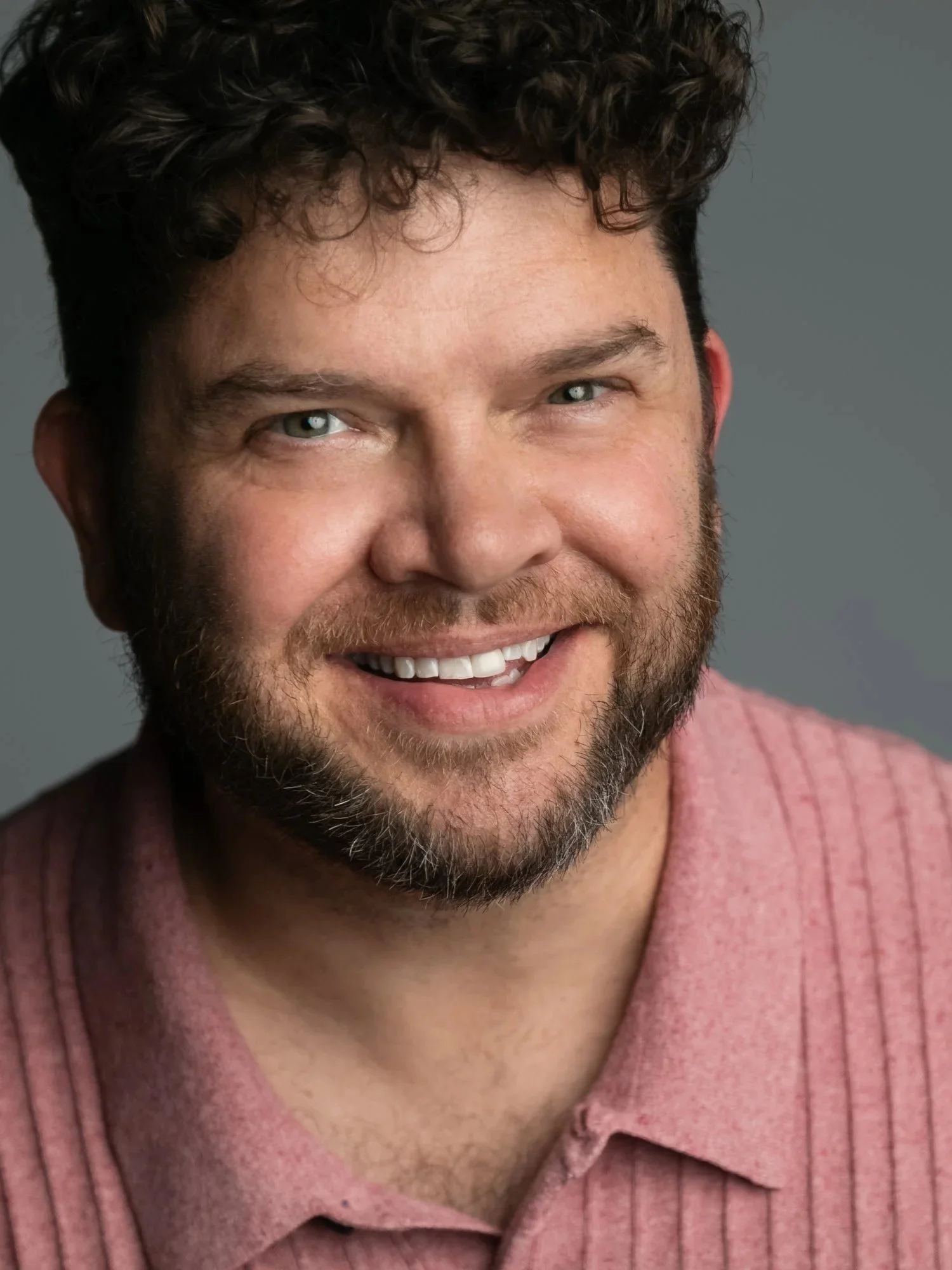 A close-up portrait of Jeremy with short, curly dark hair, and a pink striped shirt against a gray background.