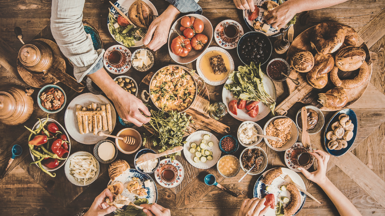 A top-down view of a family or group gathering around a wooden table filled with various Middle Eastern or Mediterranean dishes, including bread, salads, olives, and desserts, with several people reaching for food and drinks.