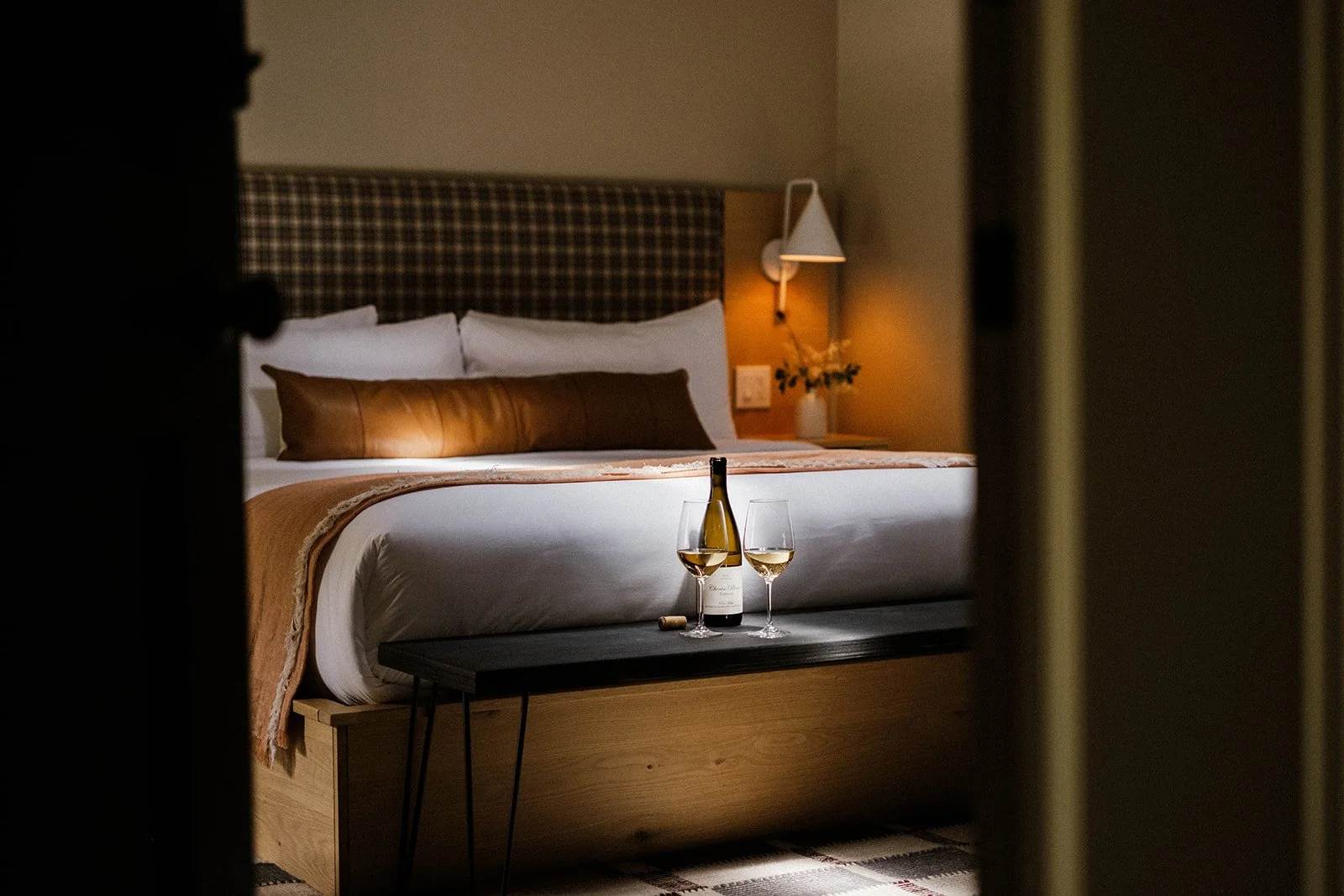 A bedroom seen through a slightly opened door, featuring a bed with white linens and a brown leather pillow, a side table with a wine bottle and two glasses, a wall-mounted lamp and a lamp on the bedside table, creating a cozy ambiance.