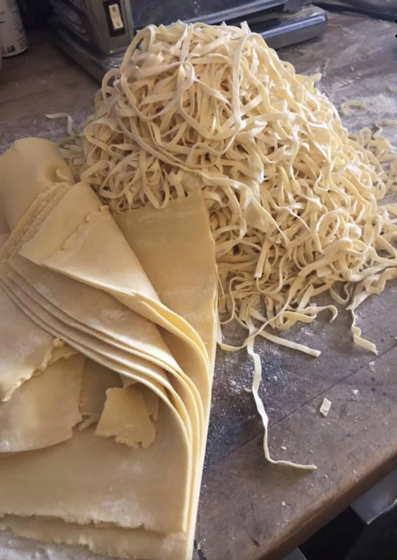A pile of fresh, uncooked fettuccine pasta on a wooden work surface, with some pasta sheets rolled and partially peeled back.