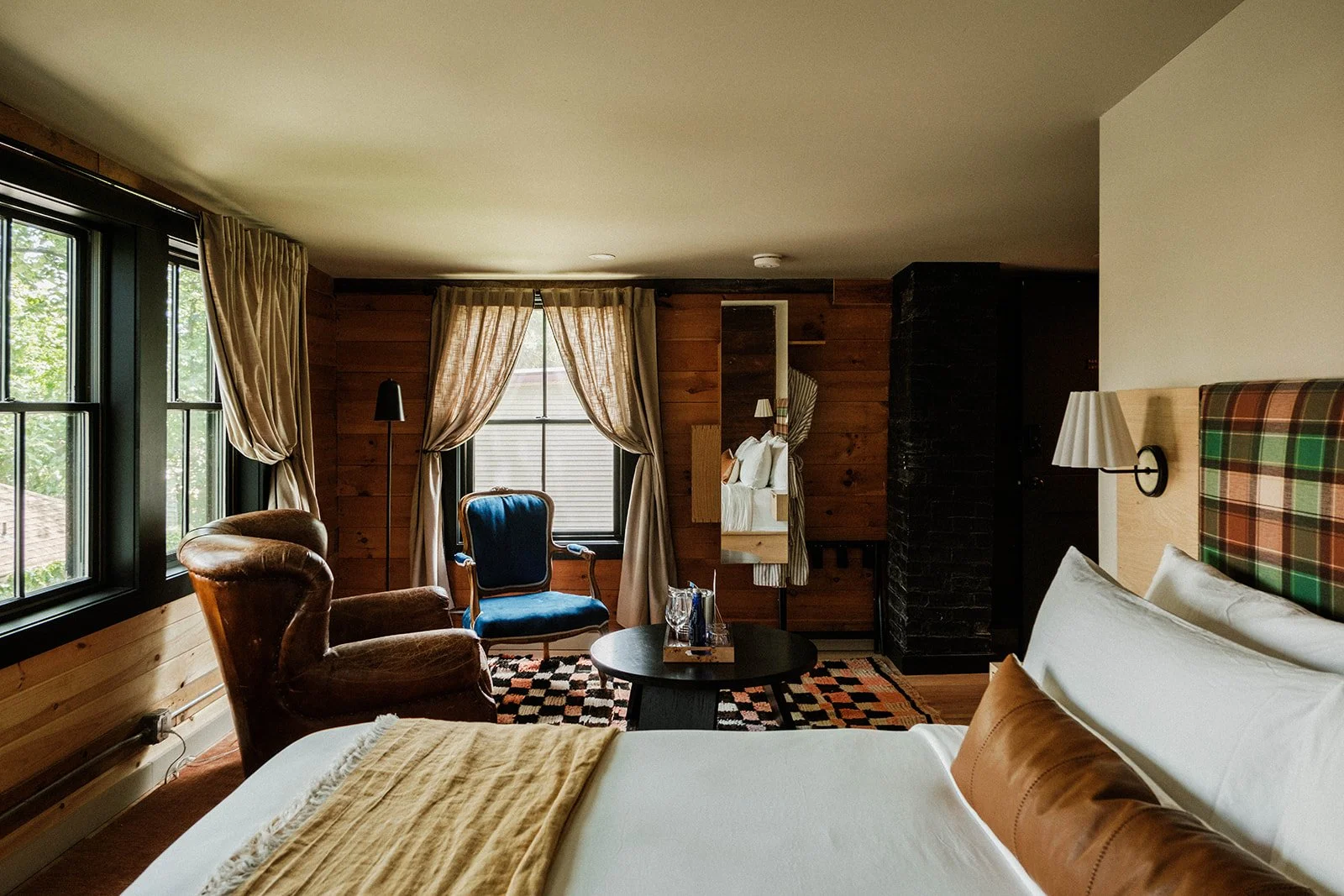 Interior of a cozy bedroom with a large bed, wooden walls, armchairs, a window with curtains, and a small round table.